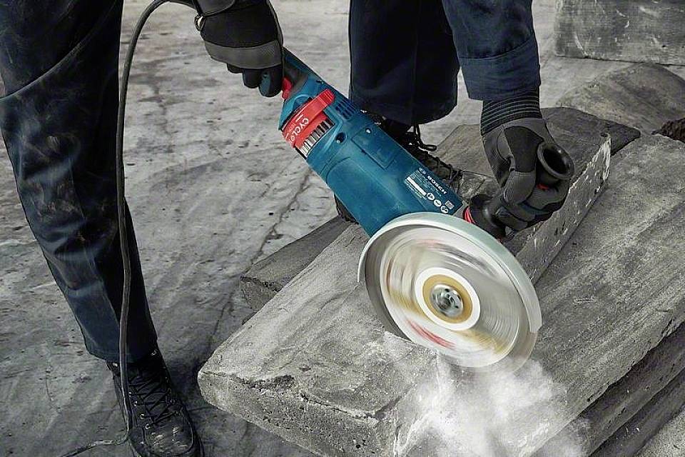 Een persoon snijdt een betonblok met een elektrische haakse slijper. Vonken spatten rond, en stof wordt opgewaaid.