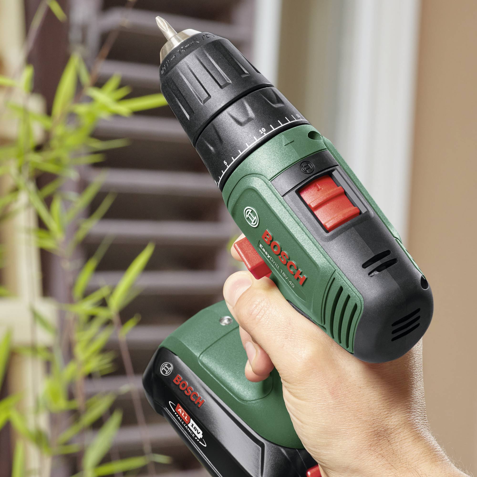 Een hand houdt een Bosch-boormachine vast, klaar om gaten te boren. Op de achtergrond zijn onscherpe planten te zien.