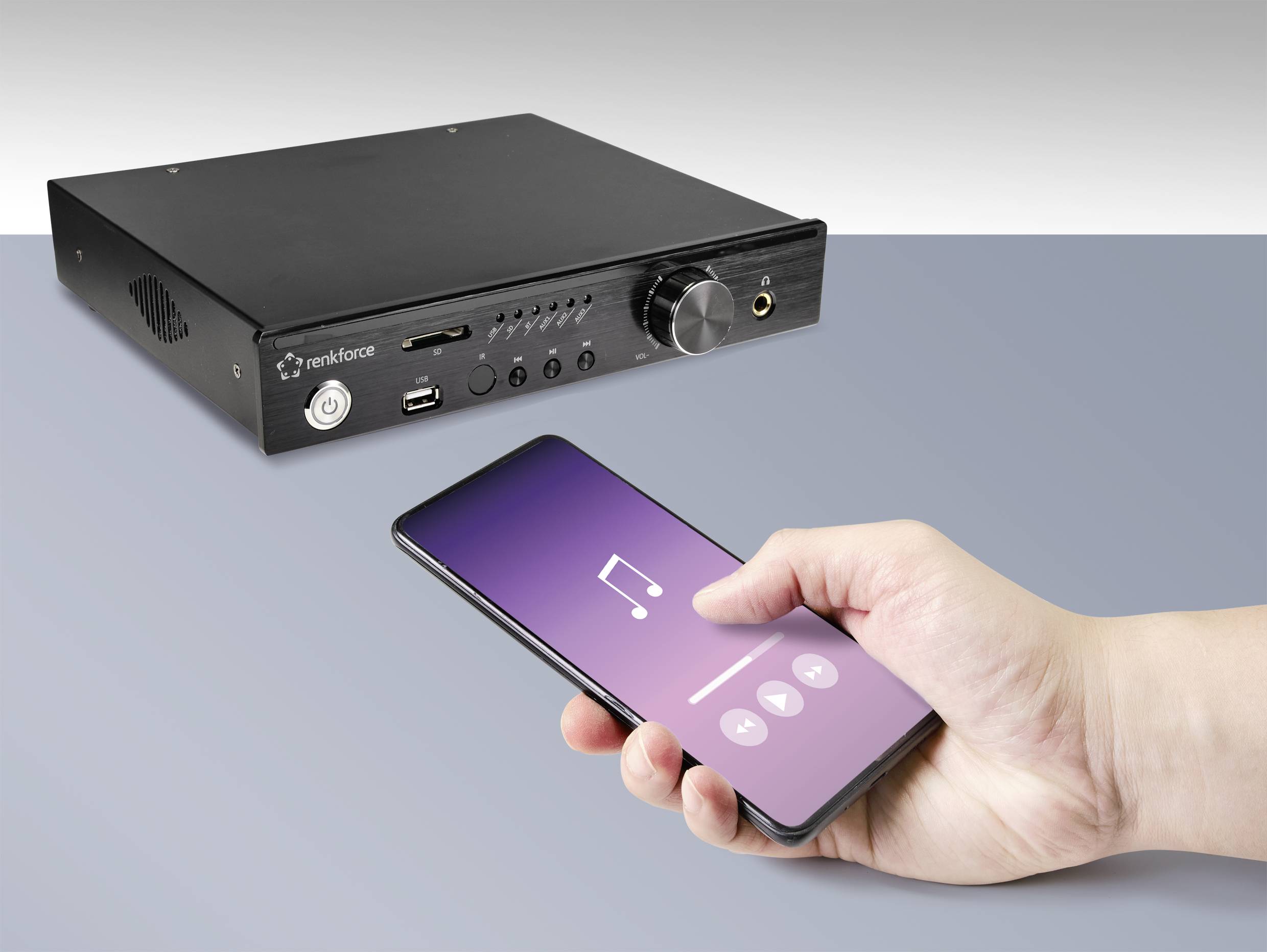 Een hand houdt een smartphone vast met een muziek-app-interface. Op de achtergrond is een zwart audioapparaat met meerdere aansluitingen zichtbaar.