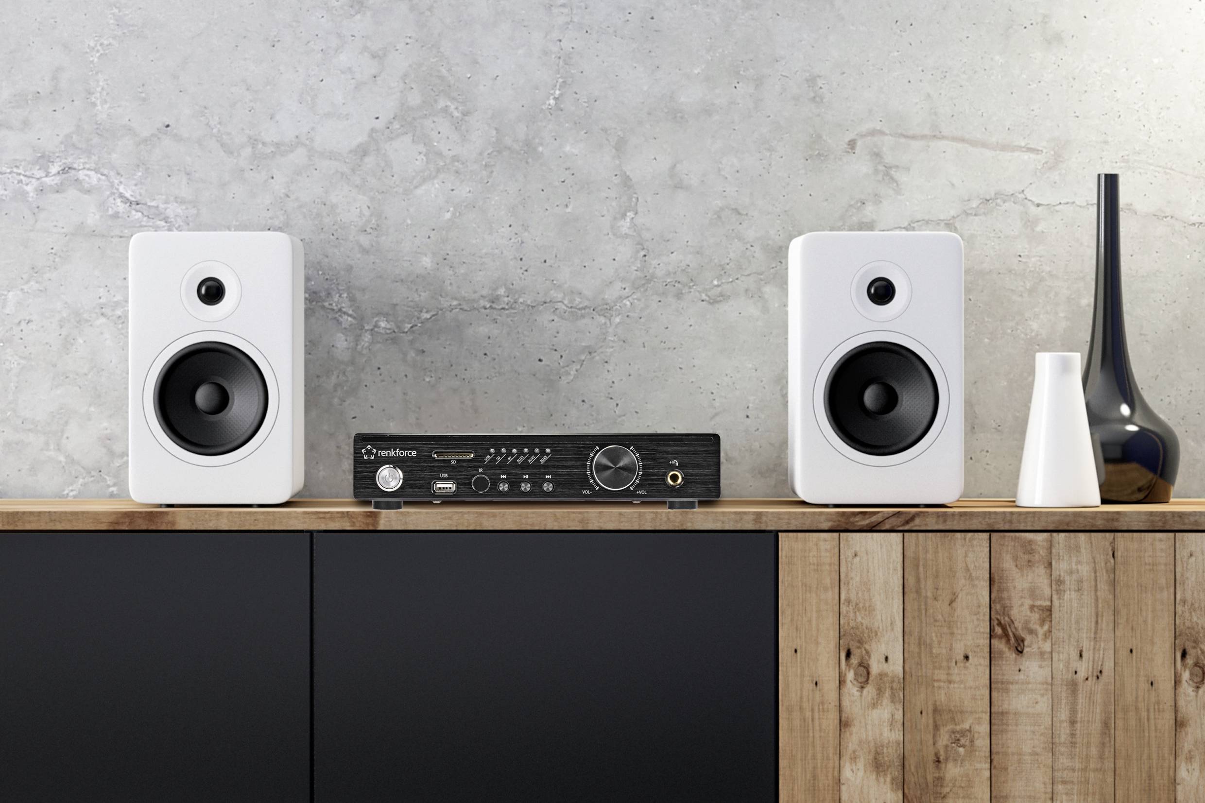 Twee witte speakers en een zwart-zilveren versterker op een houten tafel voor een grijze betonnen muur; moderne ruimte-inrichting.