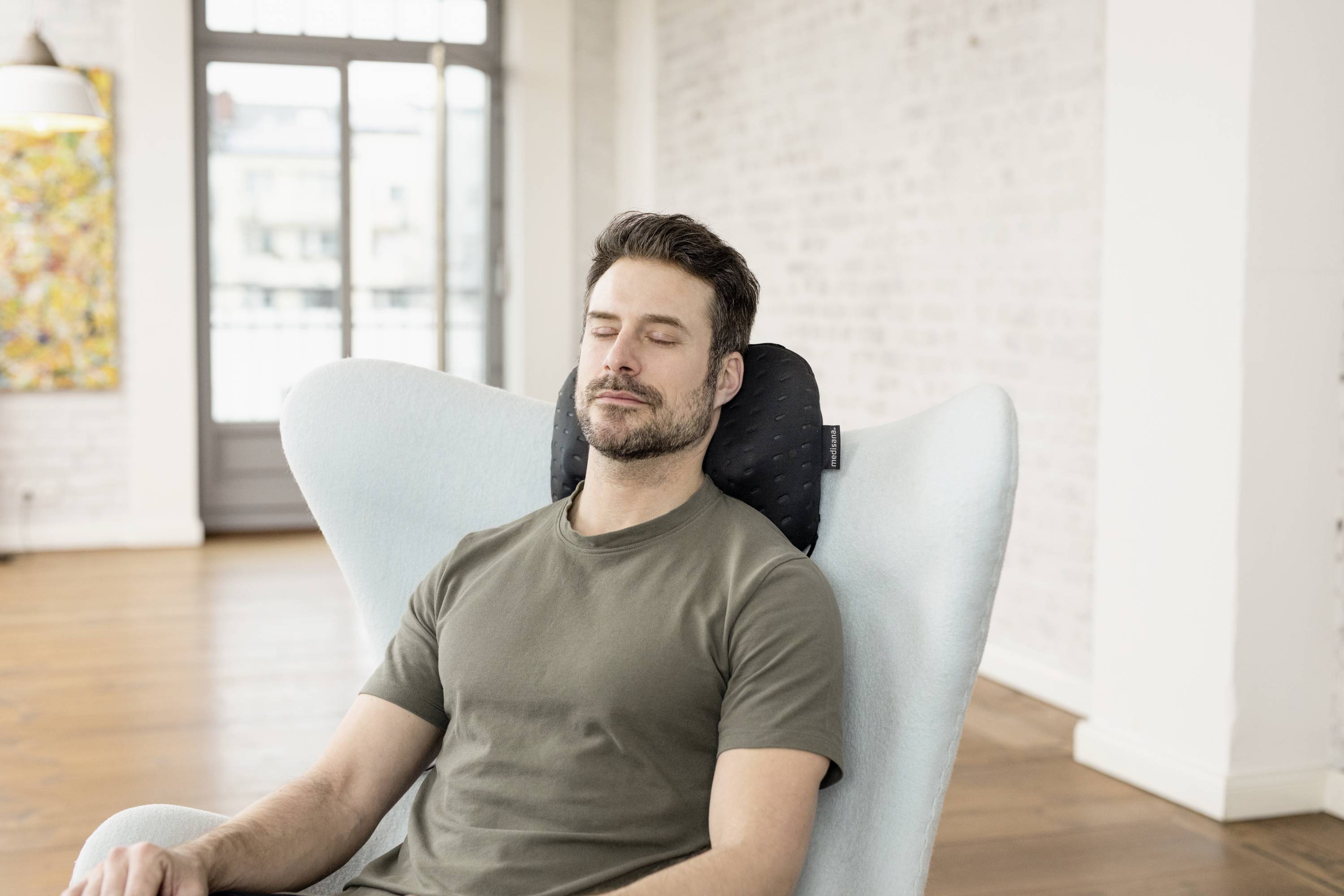 Man ontspant in een fauteuil met gesloten ogen, draagt een groen T-shirt. De achtergrond toont een lichte woonkamer met grote ramen.