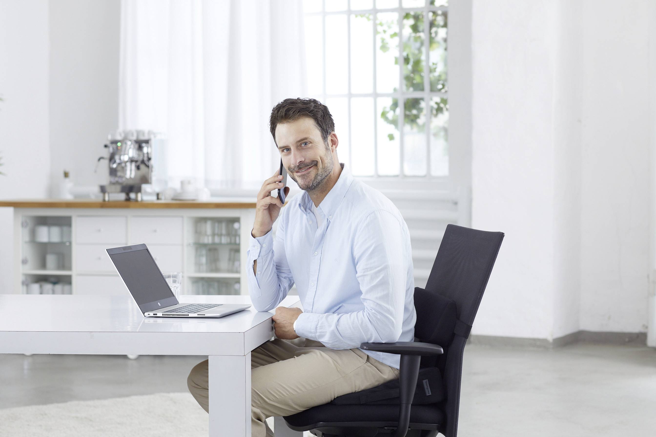Een man zit aan een tafel met een laptop, belt en glimlacht. Op de achtergrond is een licht kantoor met grote ramen te zien.