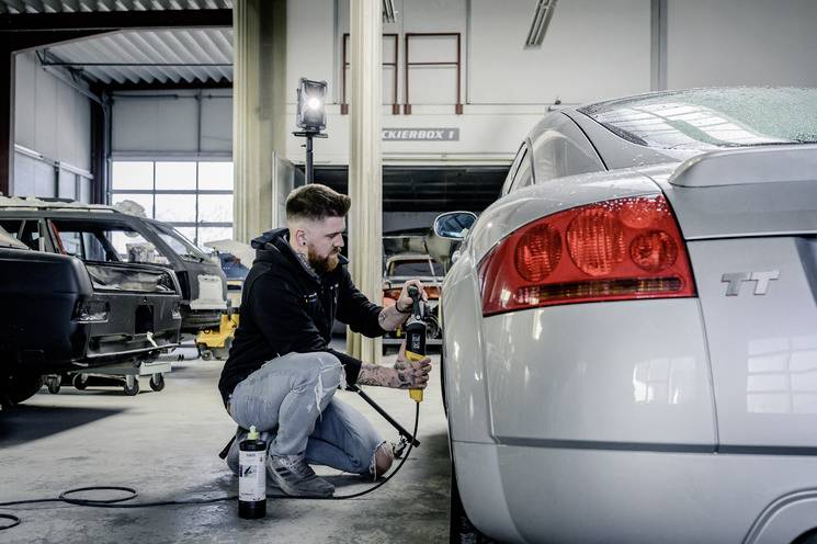 Een man werkt in een autogarage aan een zilveren sportwagen. Hij gebruikt een gereedschap om aan het voertuig te werken. Op de achtergrond staan andere auto's.