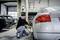 Een man werkt in een autogarage aan een zilveren sportwagen. Hij gebruikt een gereedschap om aan het voertuig te werken. Op de achtergrond staan andere auto's.