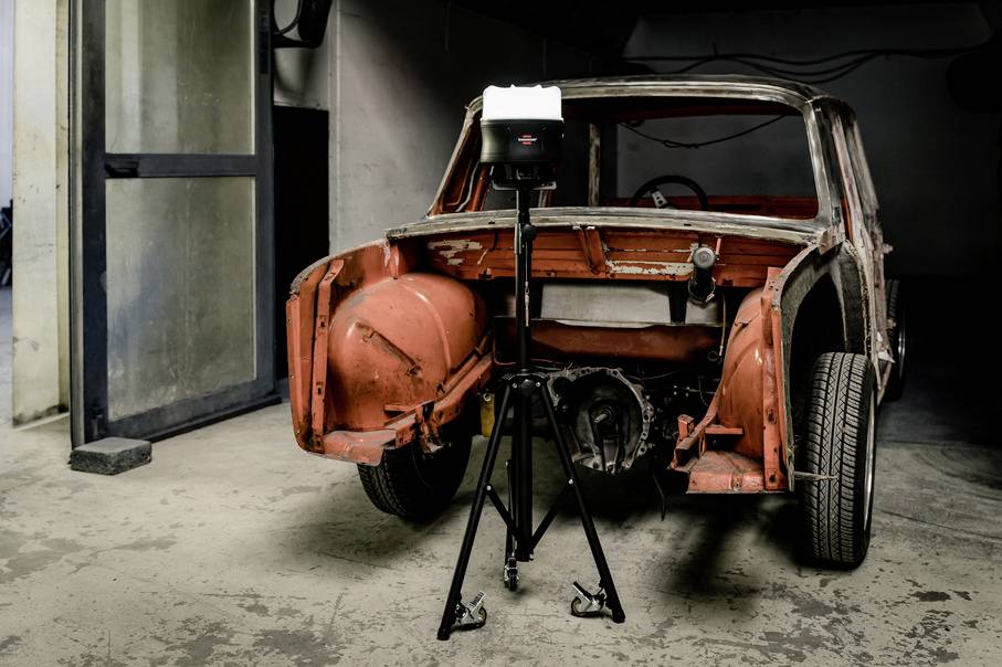 Een gedeeltelijk gerestaureerde auto zonder carrosserie staat in een werkplaats. Een statief met verlichting staat ervoor om het voertuig te belichten.