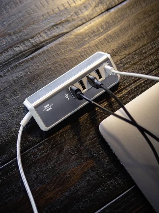 Een USB-hub met meerdere aangesloten kabels op een houten tafel. Naast de hub is een deel van een laptop zichtbaar.
