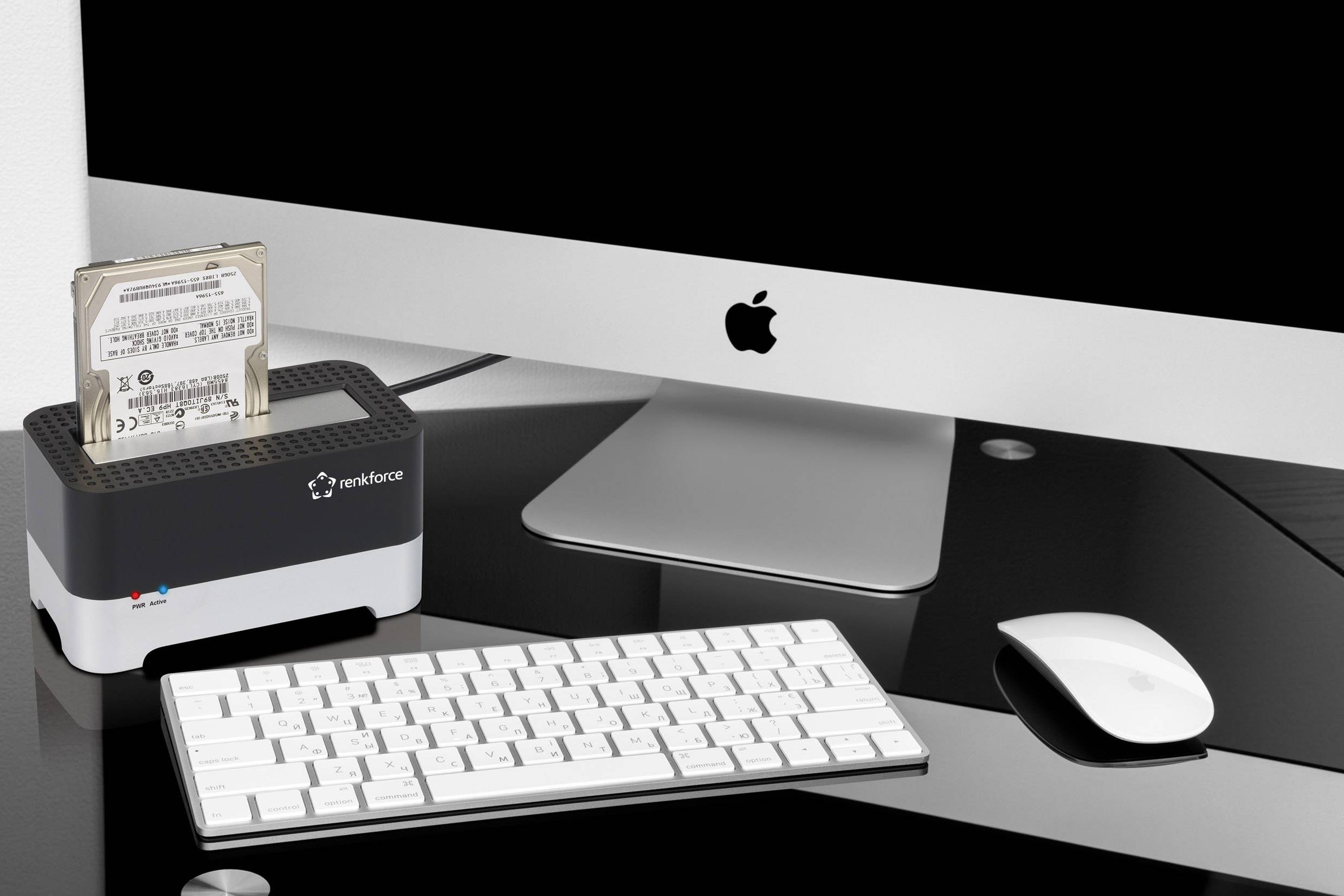 Werkplek met een iMac, externe harde schijfdock, toetsenbord en muis op een zwarte glazen tafel. Scherm toont een zwarte achtergrond.