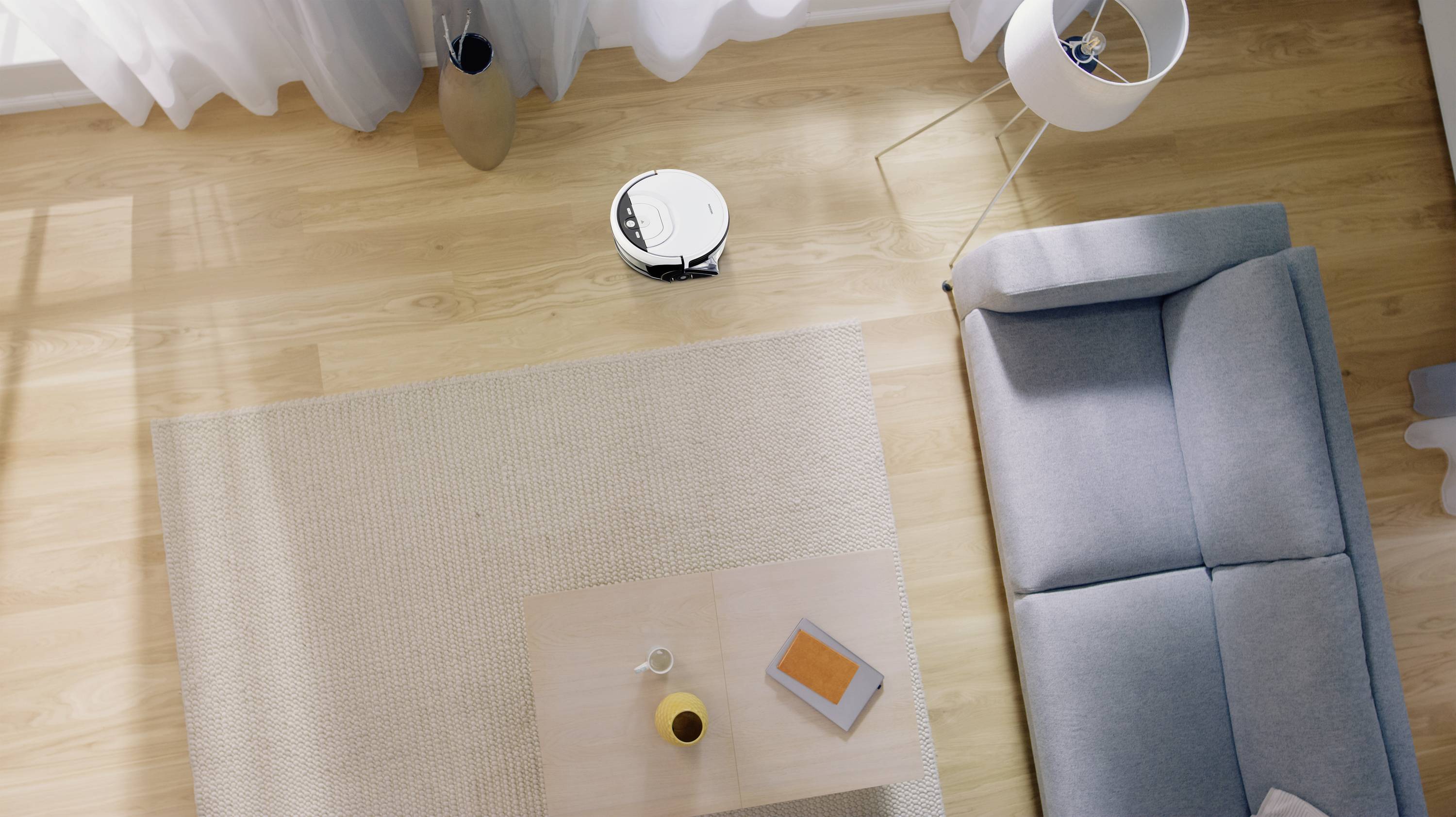 Een robotstofzuiger reinigt de lichte houten vloer van een modern woonvertrek naast een bank en een salontafel met boek en vaas.