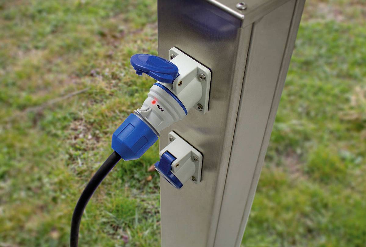 Een metalen oplaadstation staat buiten, met twee stopcontacten. Eén stopcontact is in gebruik met een blauw-wit snoer dat is aangesloten.