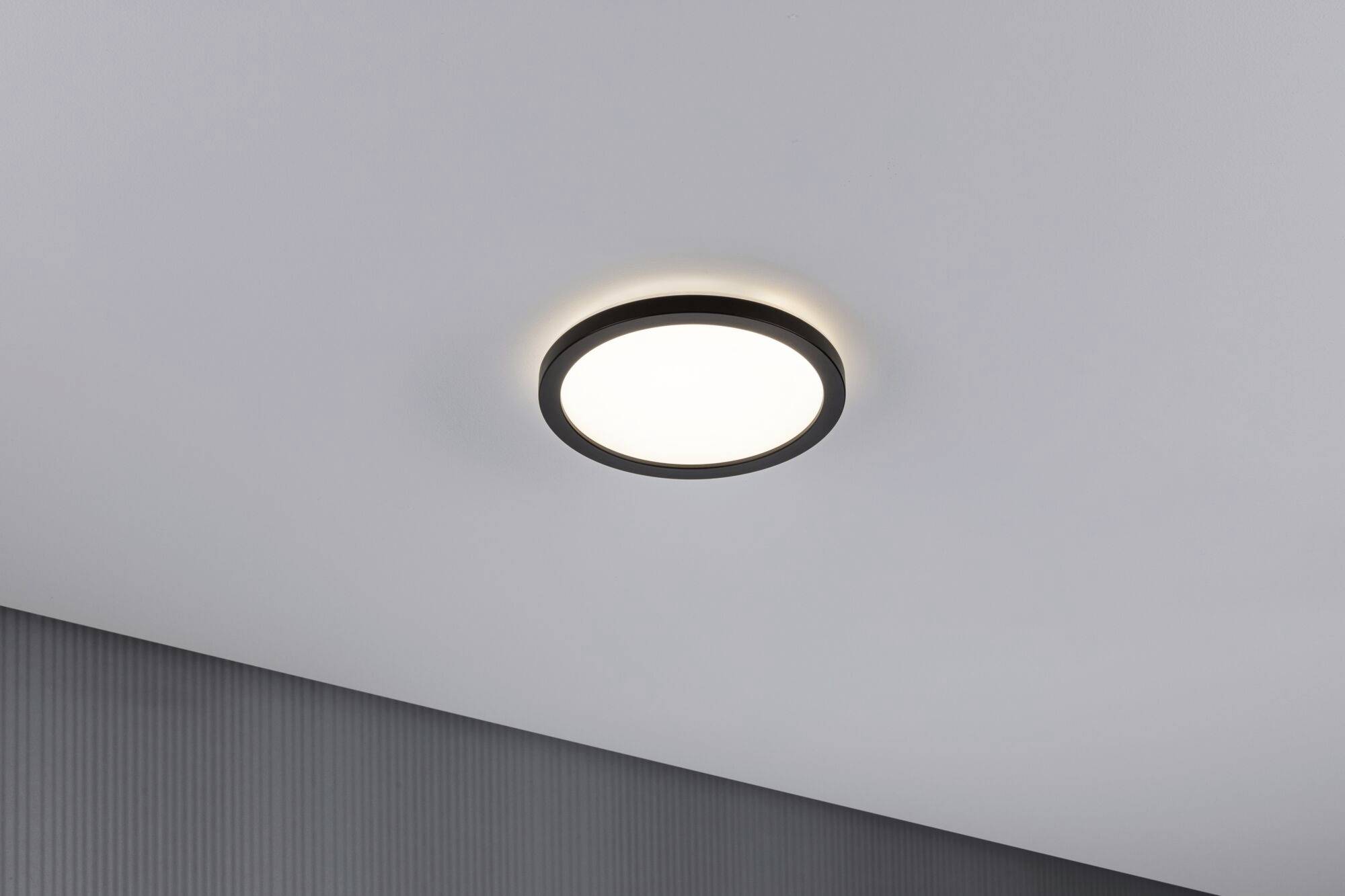 Ronde plafondlamp in een kamer, ingeschakeld en gelijkmatig schijnend, gemonteerd aan een witte plafond.