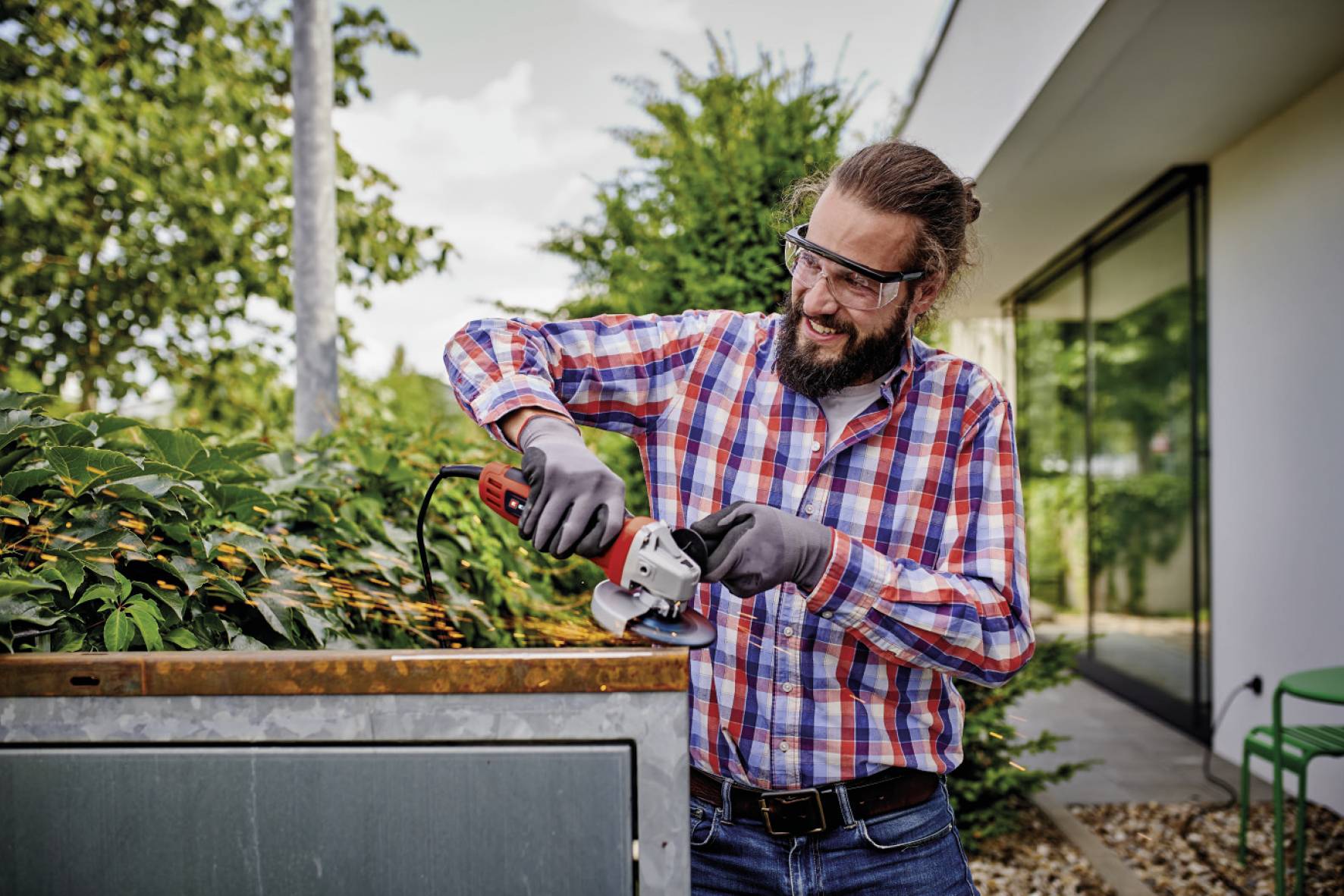 Een man met een baard en veiligheidsbril knipt met een elektrische heggenschaar de toppen van een haag voor een modern gebouw.