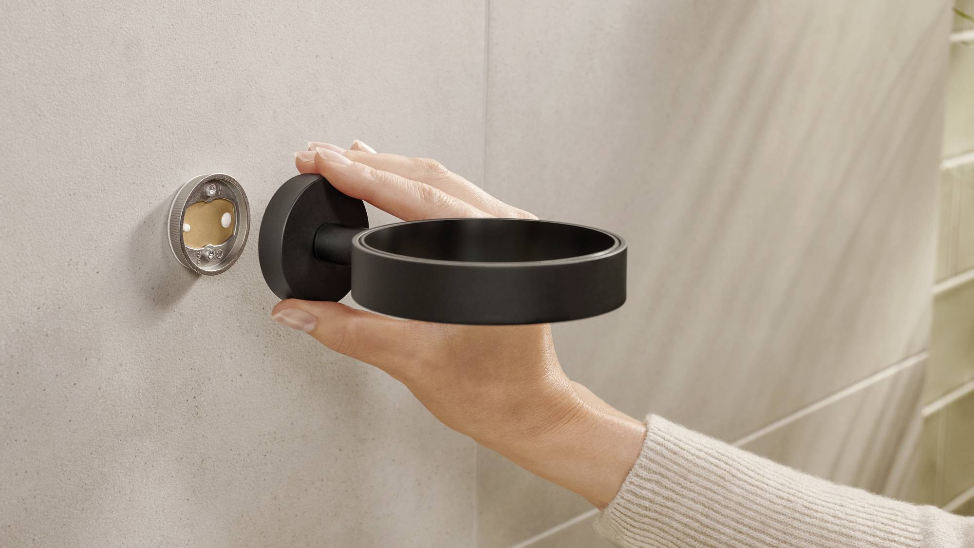 Een hand bevestigt een zwarte houder aan een grijze muur, vermoedelijk voor een badkameraccessoire zoals een beker of zeepdispenser.