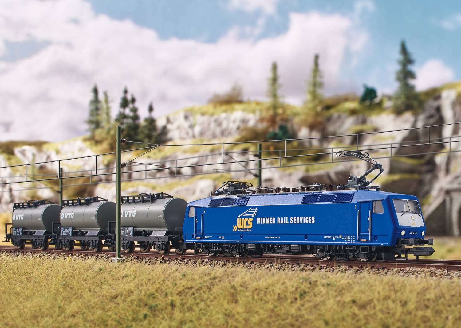 Een modeltreintje met een blauwe locomotief trekt drie zwarte tankwagons langs een landschapsachtergrond met bomen en rotsen.