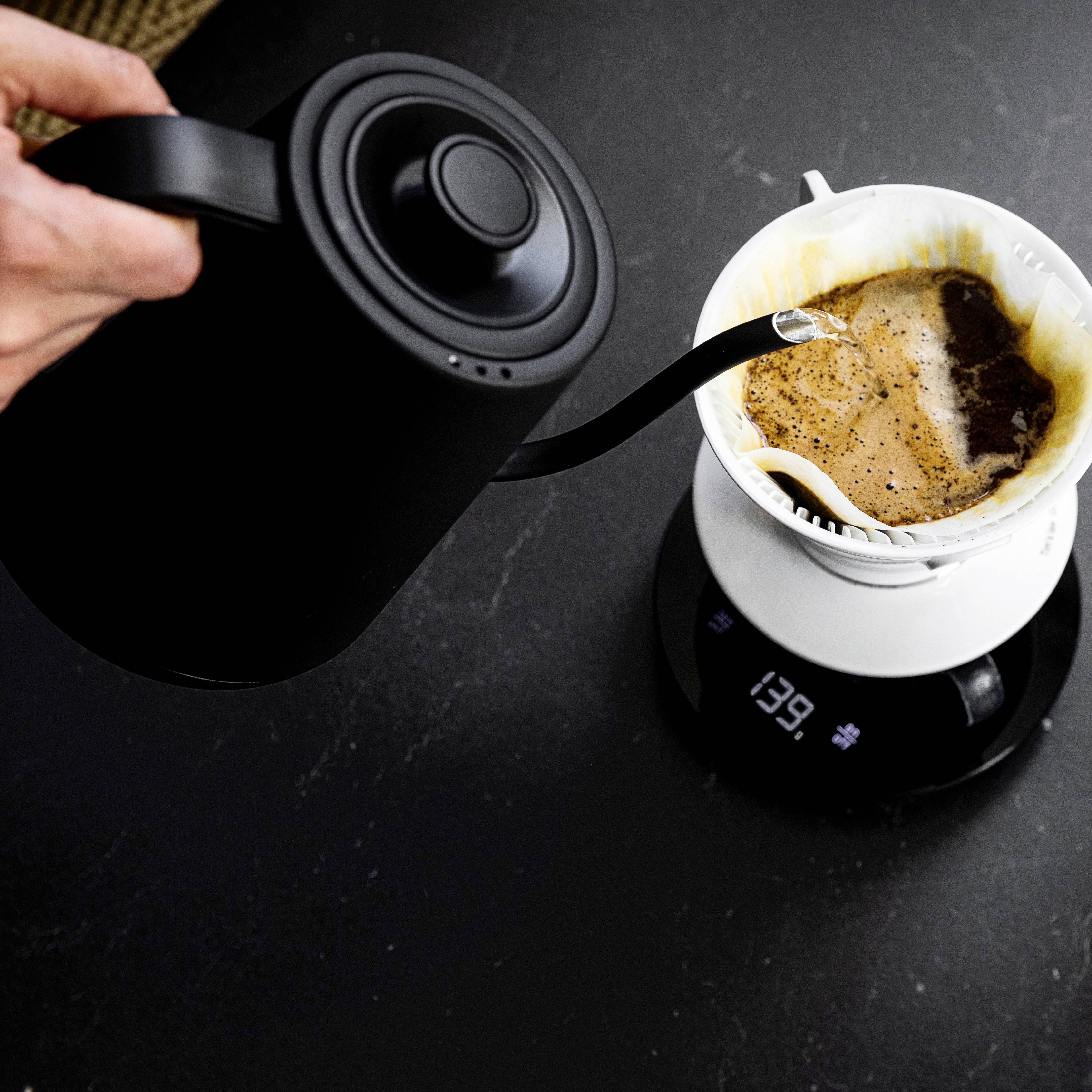 Een hand giet water uit een zwarte waterkoker in een witte koffiefilter op een weegschaal. Stoom stijgt op.