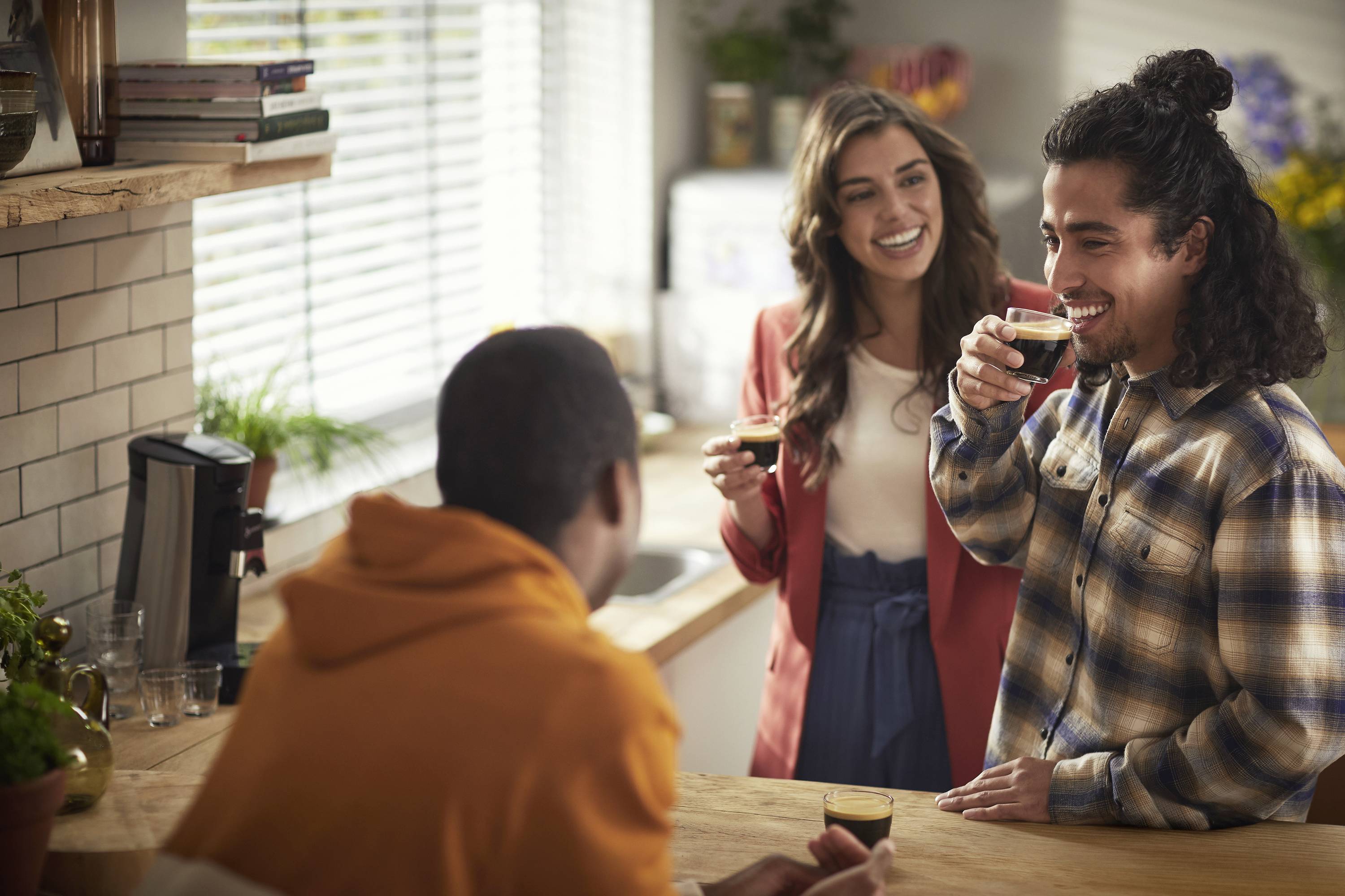 Drie personen lachen en praten met elkaar onder het genot van een kopje koffie in een gezellige keuken.