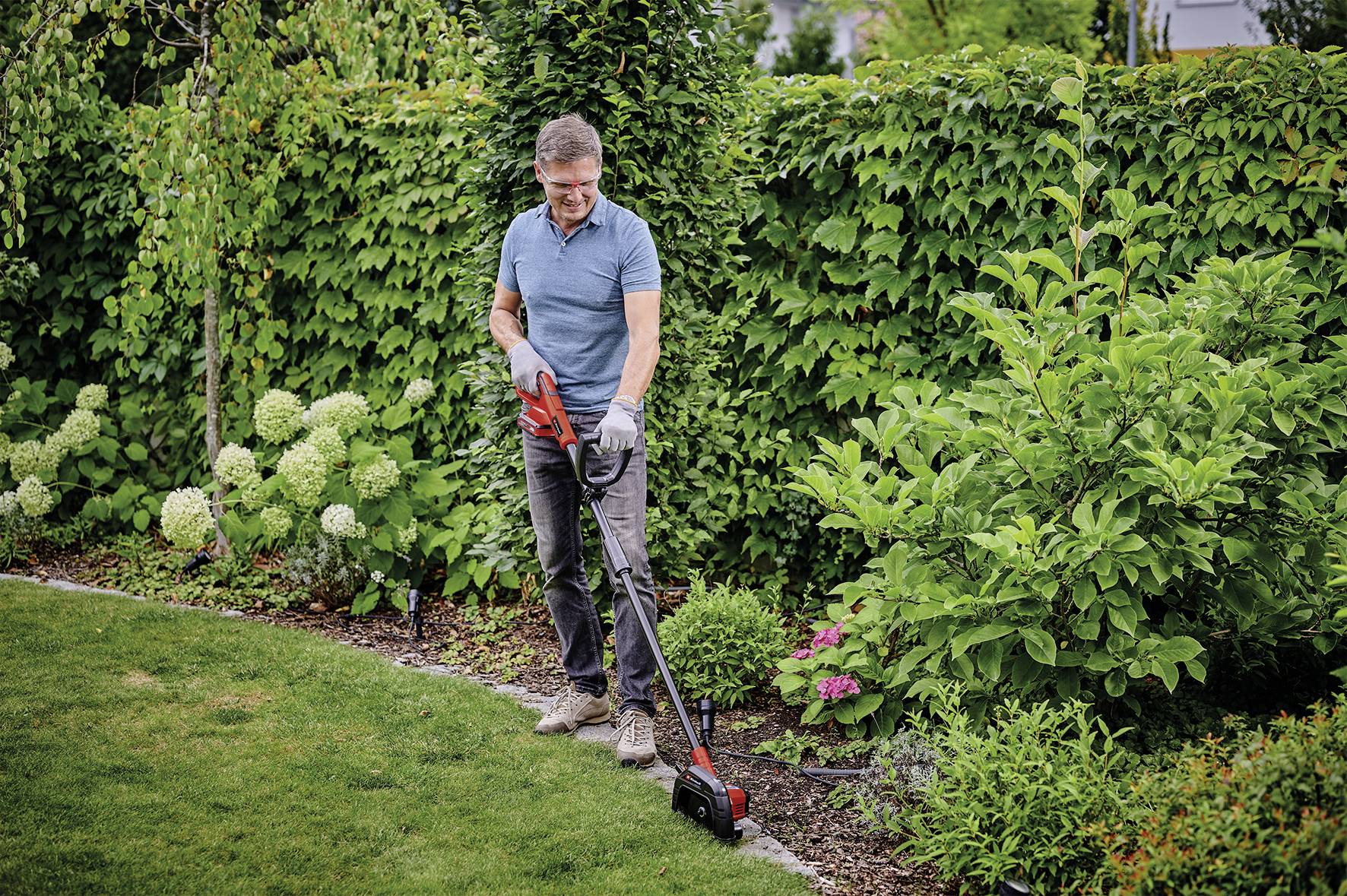 Een man maait met een elektrische trimmer de graskant in een goed onderhouden tuin met groene struiken en bloemen.