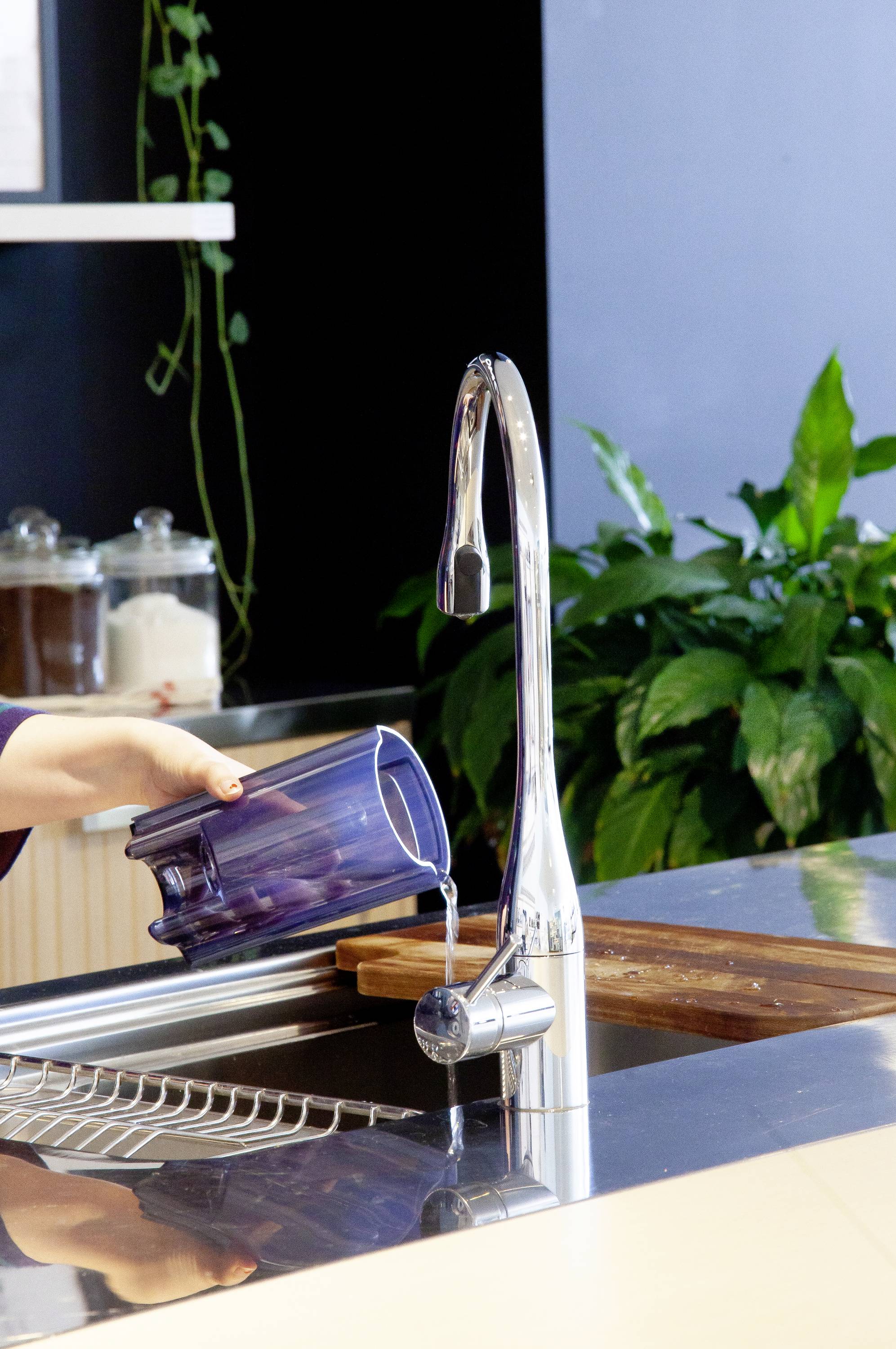 Een persoon vult een blauwe kan met water in een wasbak. Op de achtergrond zijn plantenversiering en keukengerei te zien.