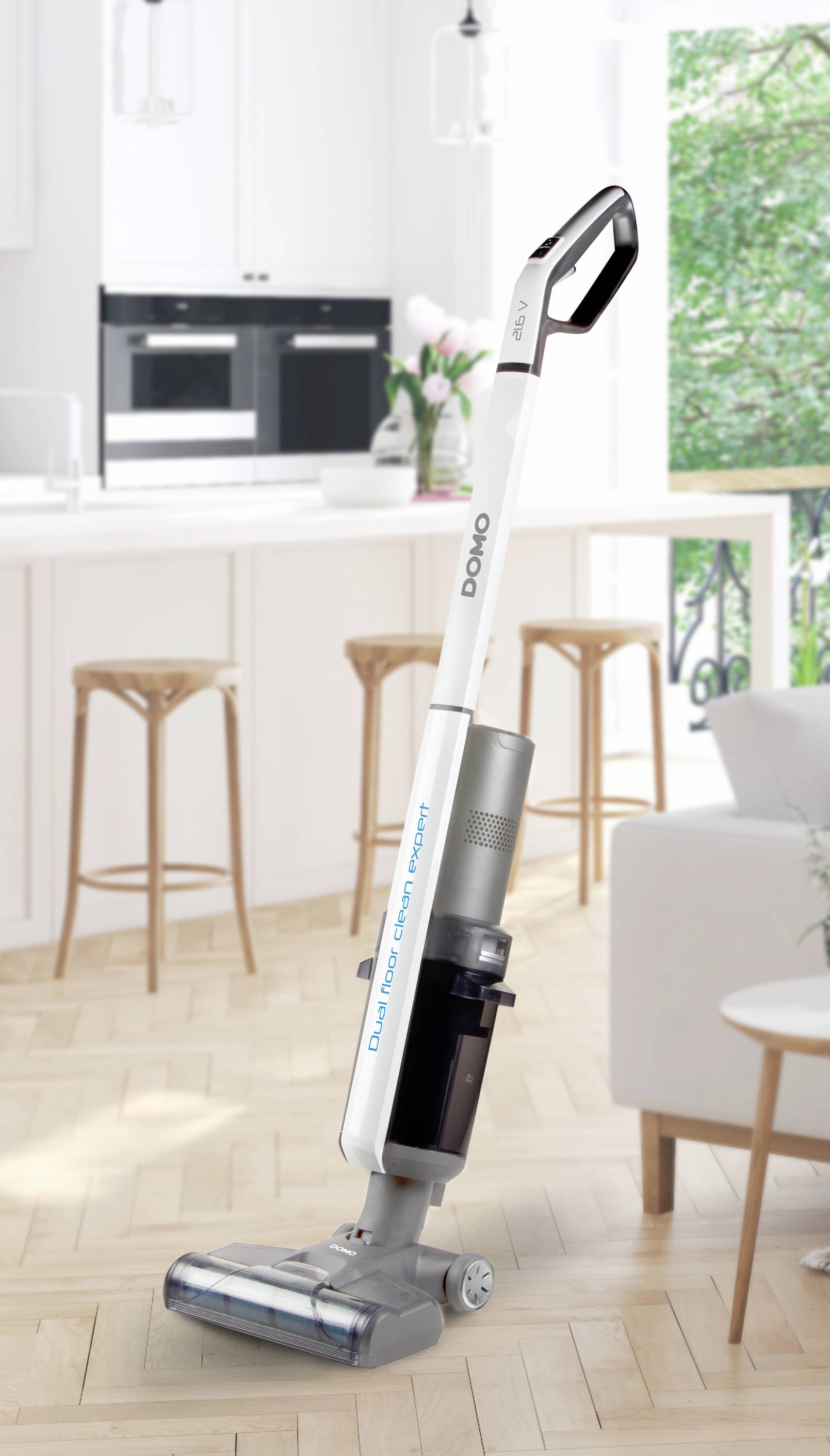 Een witte draadloze stofzuiger staat op een lichte houten vloer in een moderne keuken met witte inrichting en grote ramen.