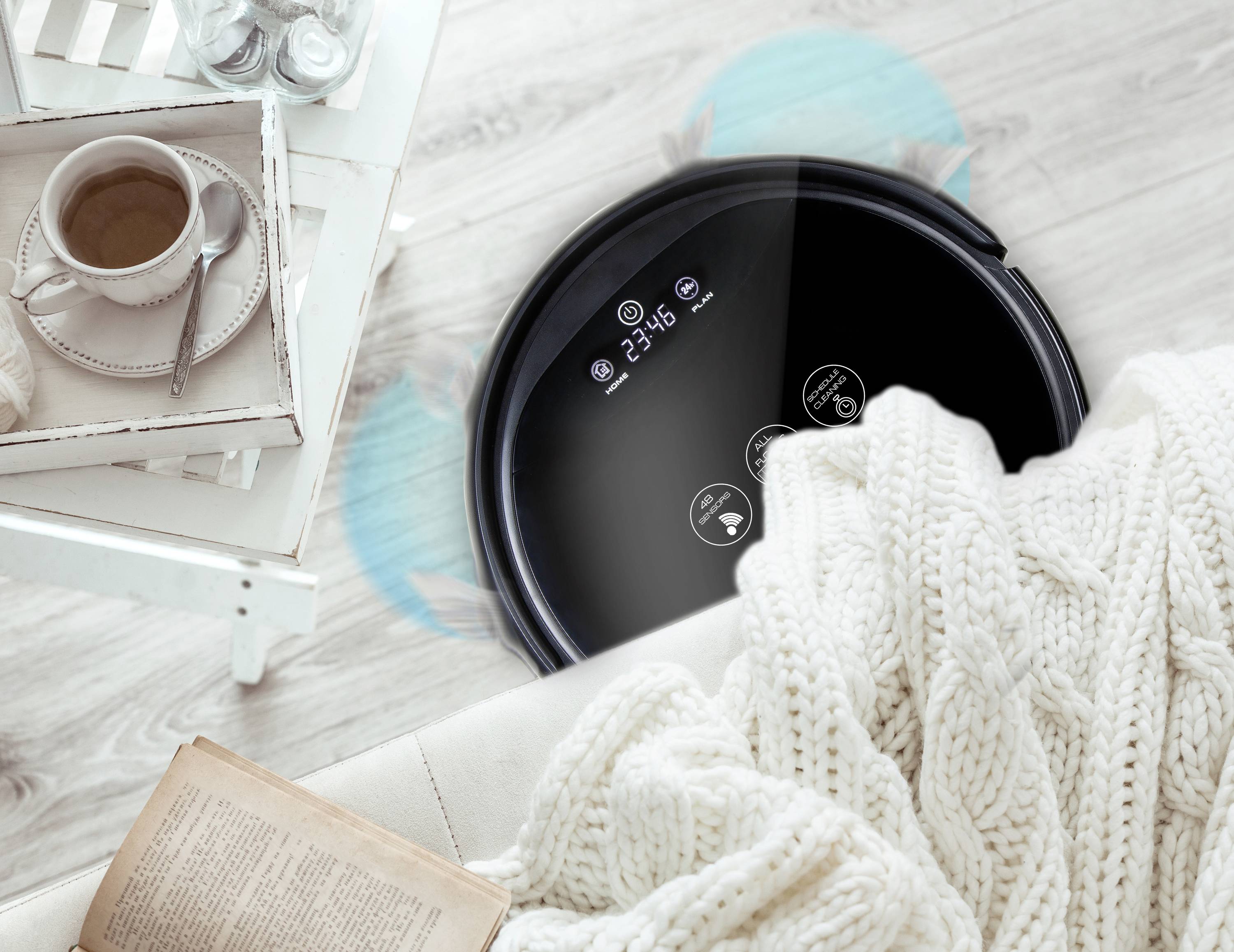 Een zwarte robotstofzuiger reinigt de vloer naast een tafel met een kopje koffie en een opengevouwen deken op een stoel.