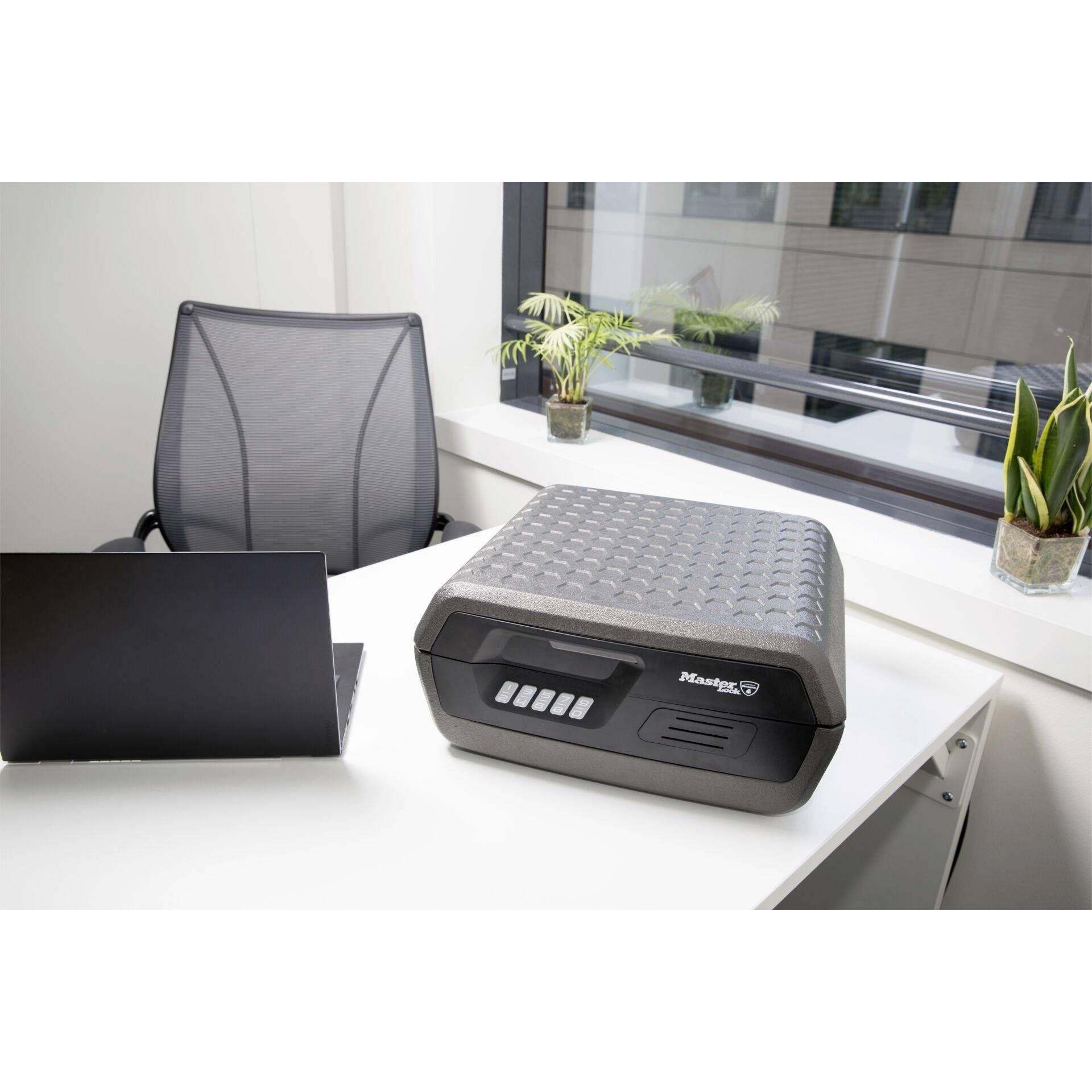 Een bureau met een zwarte laptop, een grijze archiefkast en drie planten op de vensterbank.