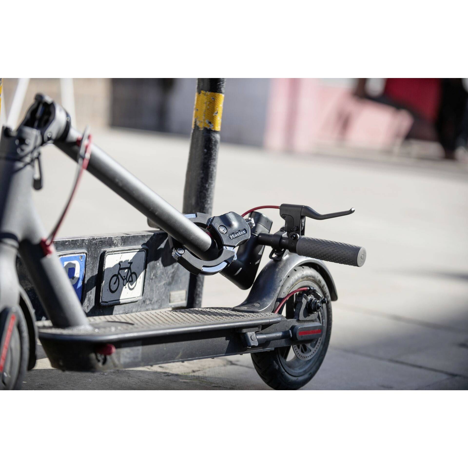 Een elektrische scooter is met een slot bevestigd aan een fietsrek. Op de achtergrond zijn wazig voetgangersgebieden zichtbaar.