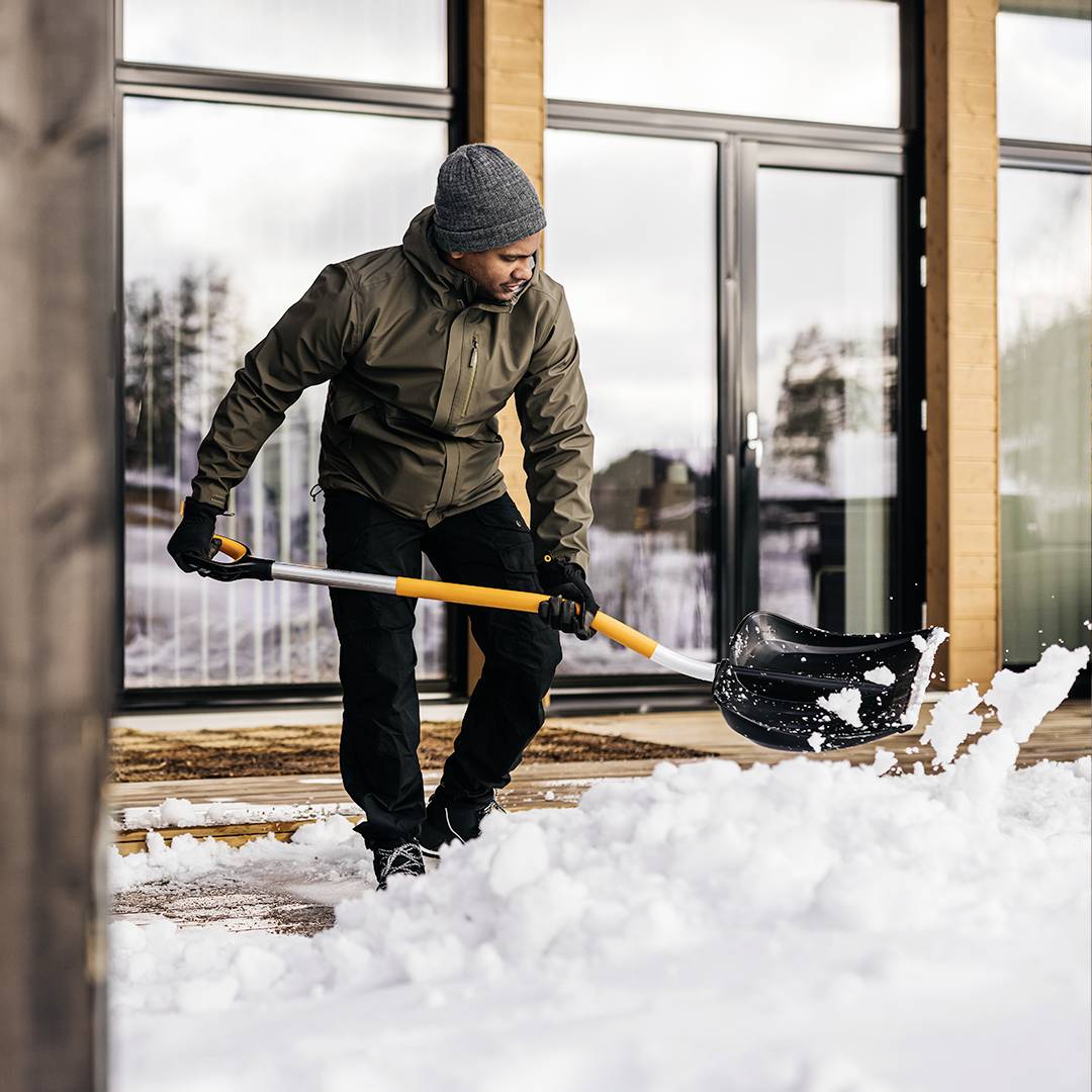Een persoon schept sneeuw voor een modern gebouw met grote ramen. Winterse scenerie, waarschijnlijk koude temperaturen.