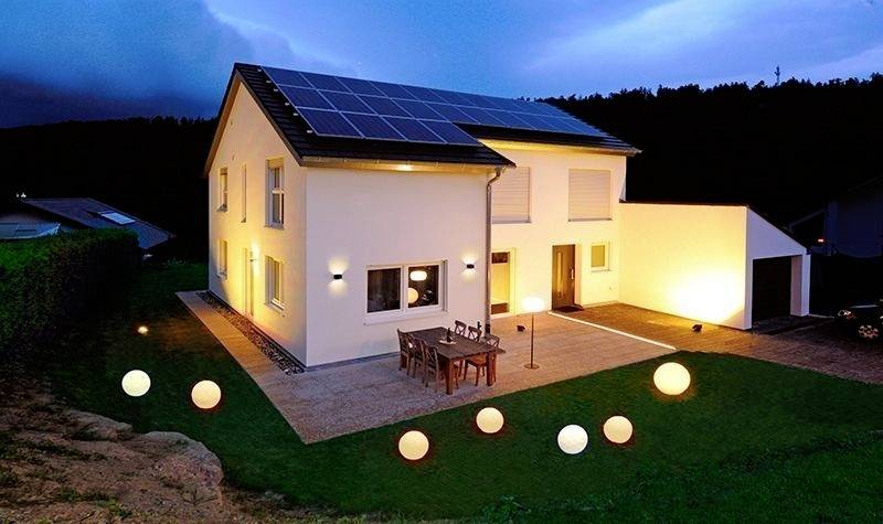 Vrijstaand huis bij schemering met zonnepanelen op het dak en verlichte tuin; terras met tafel en stoelen.