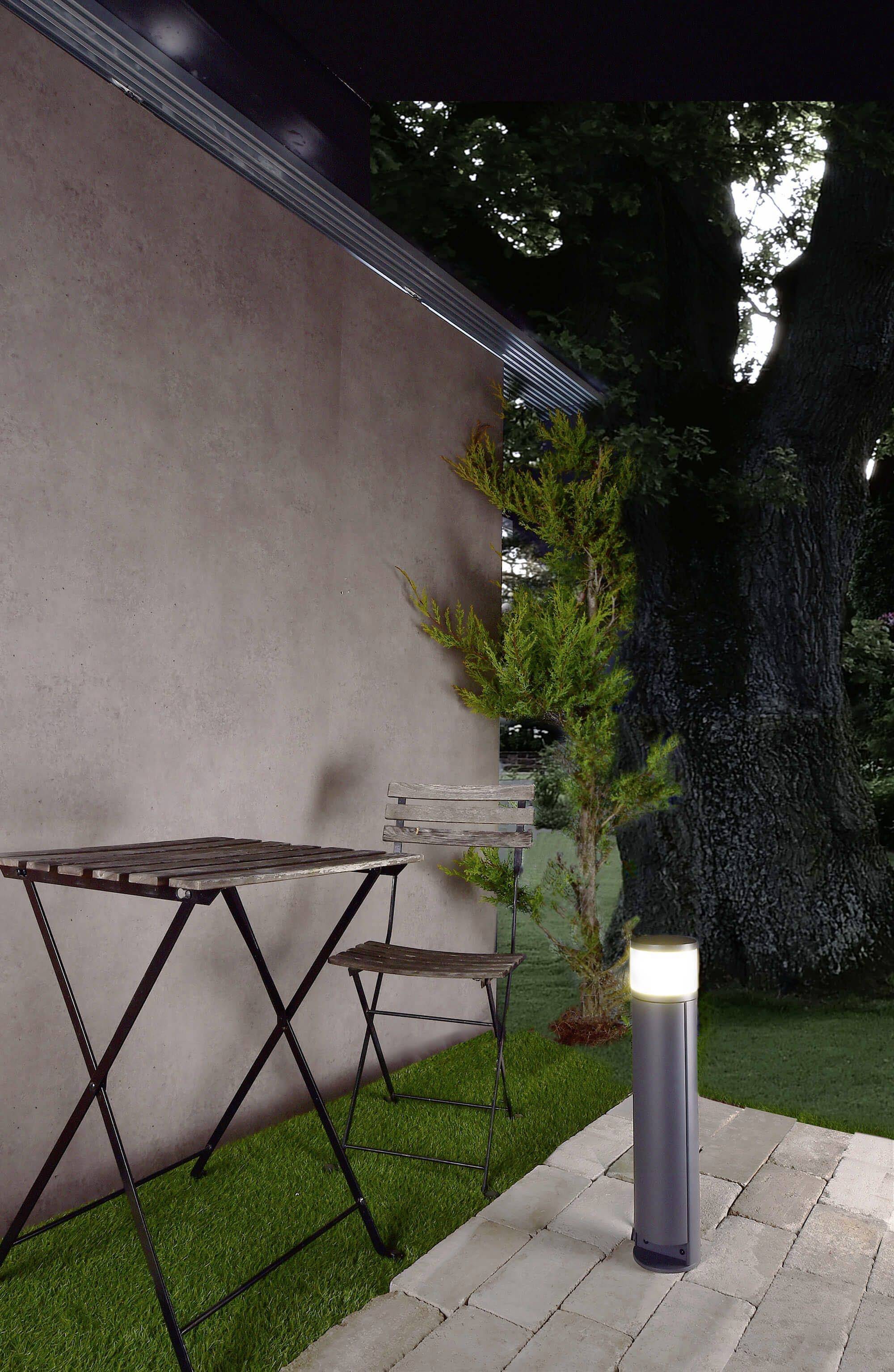 Een kleine houten tafel en stoel staan op een terras. Ernaast een moderne staande lamp en een boom op de achtergrond.