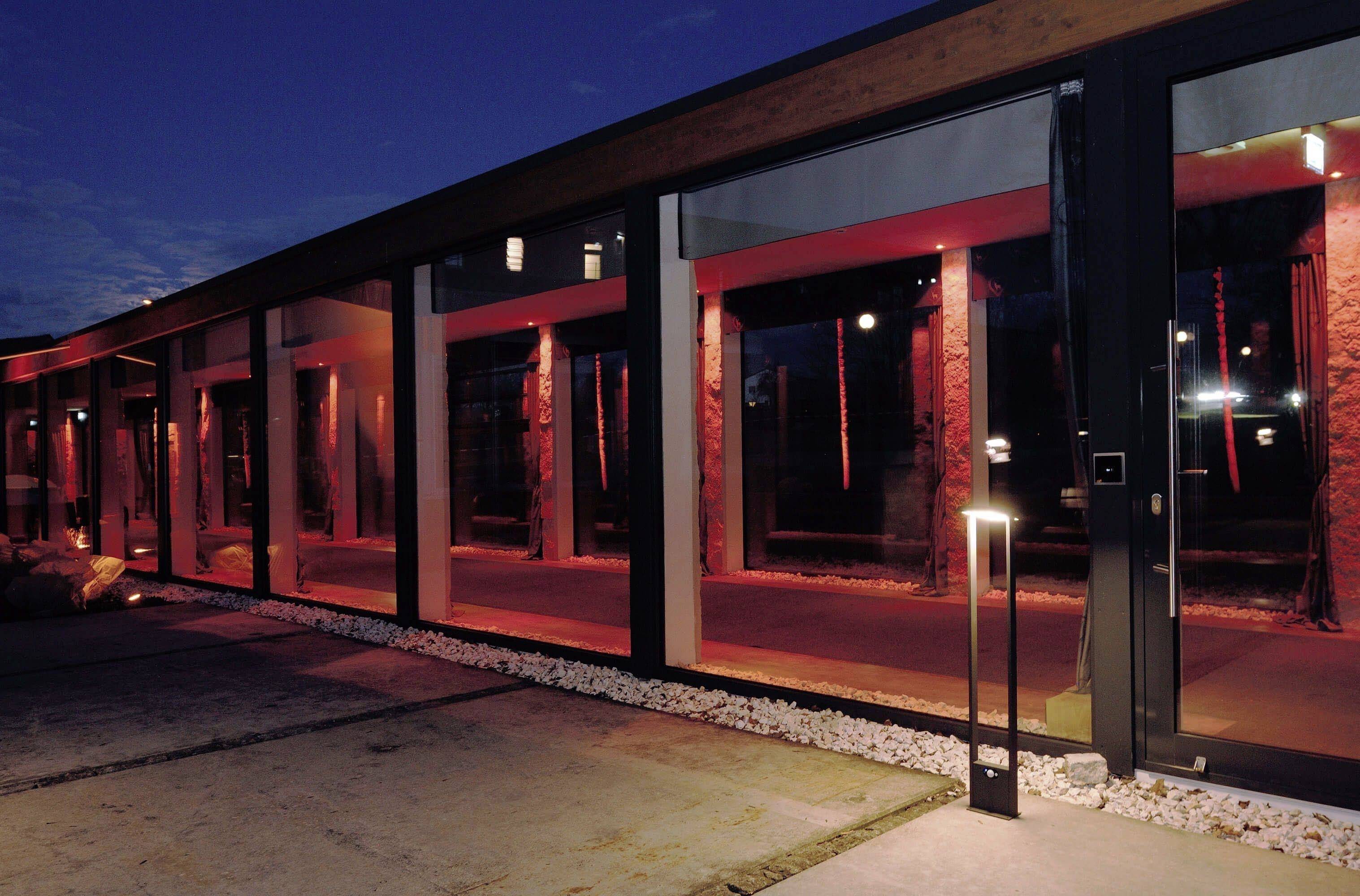 Een modern gebouw met grote ramen, verlicht door rood licht dat 's nachts een warme en uitnodigende sfeer creëert.