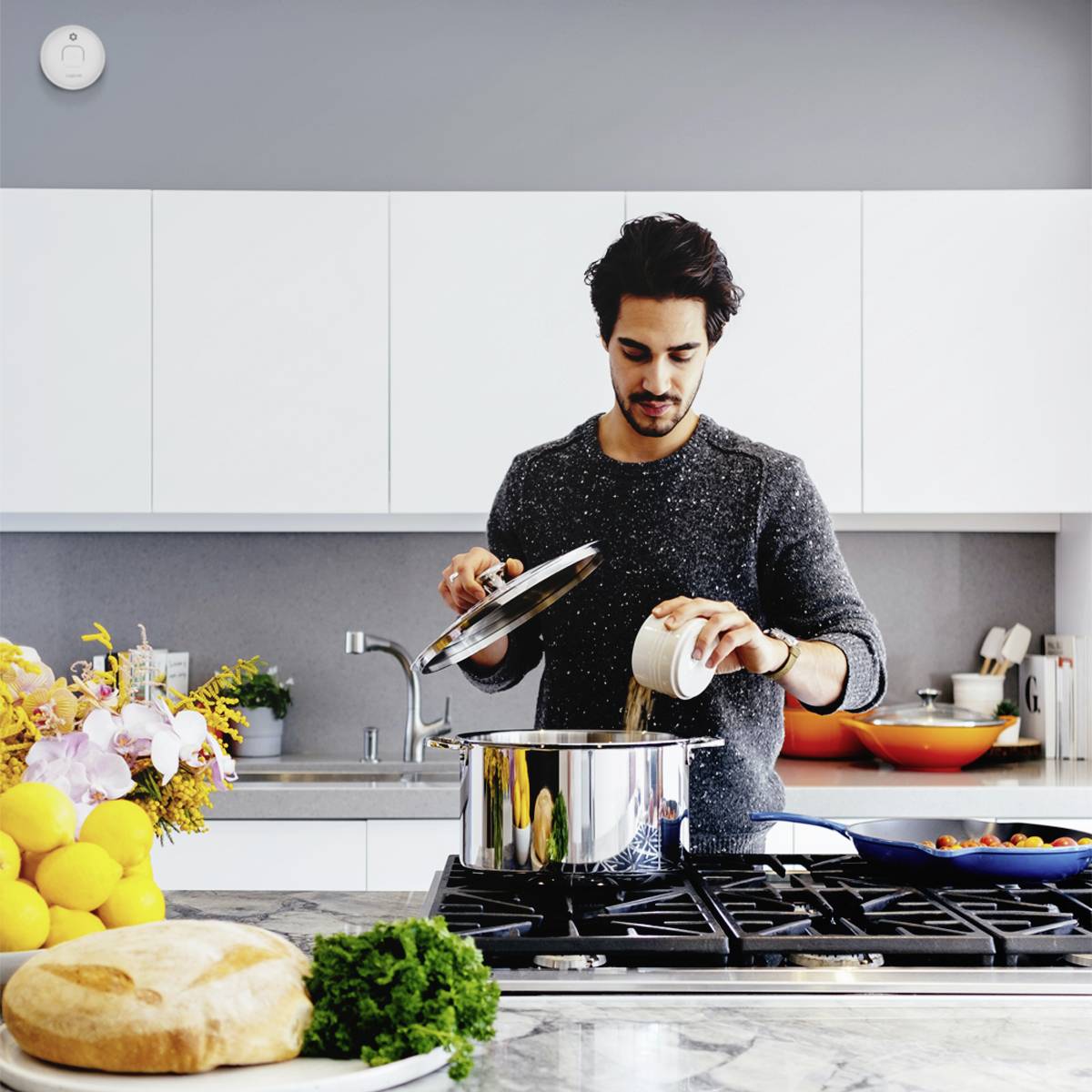 Een man kookt in een moderne keuken. Hij tilt het deksel van een pan op en giet er een vloeistof in. Op de achtergrond zijn keukenkastjes en een fornuis te zien.