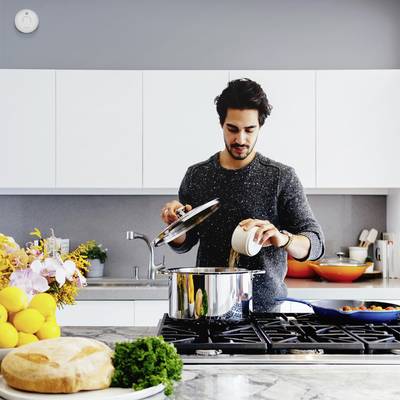 Een man kookt in een moderne keuken. Hij tilt het deksel van een pan op en giet er een vloeistof in. Op de achtergrond zijn keukenkastjes en een fornuis te zien.