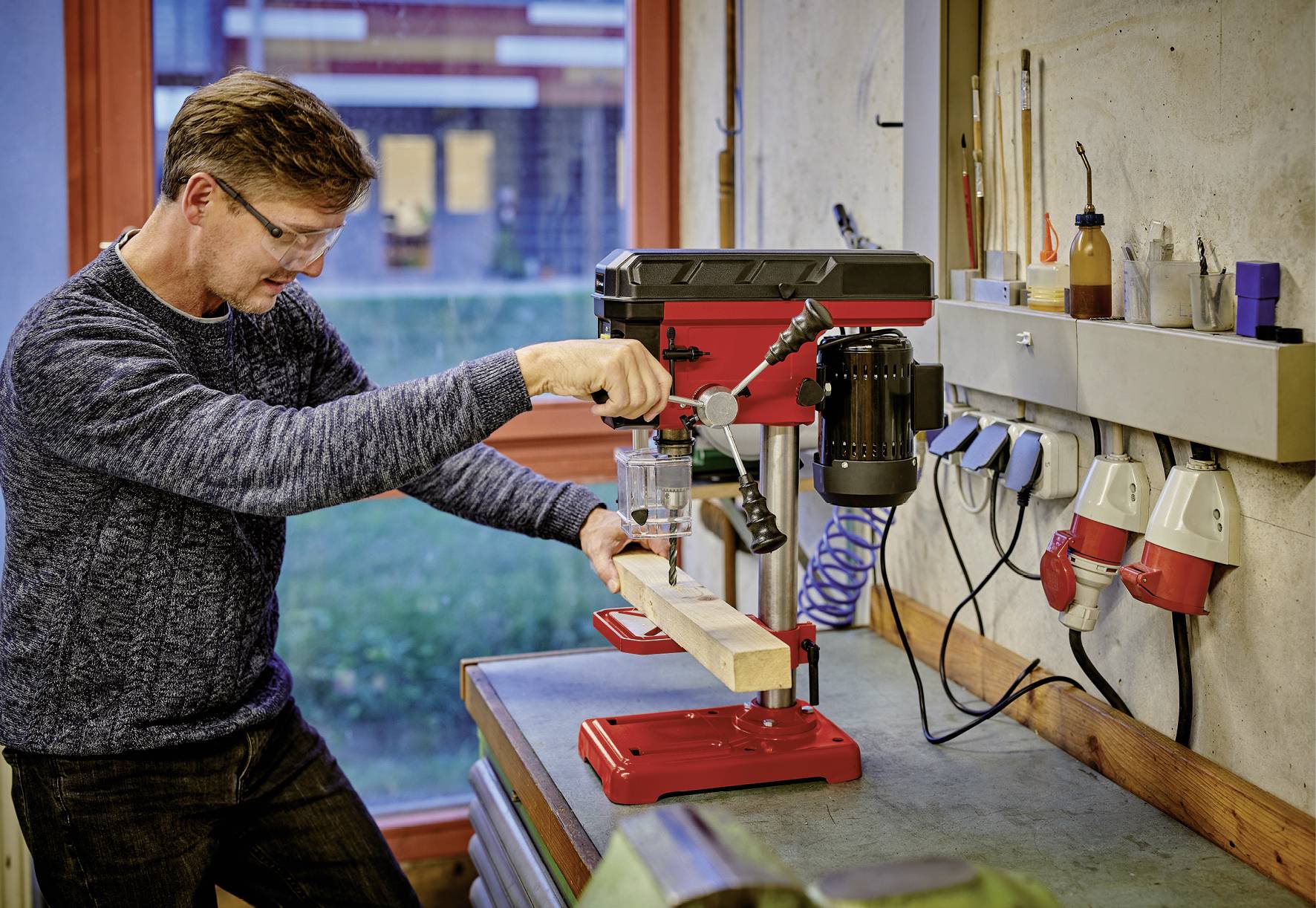 Een man met veiligheidsbril bedient een tafelboormachine in een werkplaats en boort gaten in een houten plank. Op de achtergrond zijn gereedschappen zichtbaar.