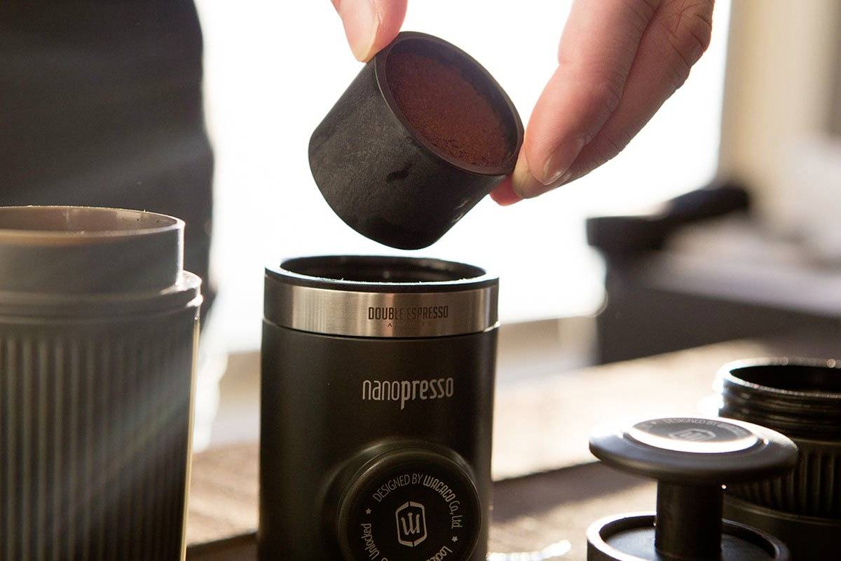 Een hand vult gemalen koffie in een draagbare espressomachine. Het is een zonnige omgeving met accessoireonderdelen op de achtergrond.