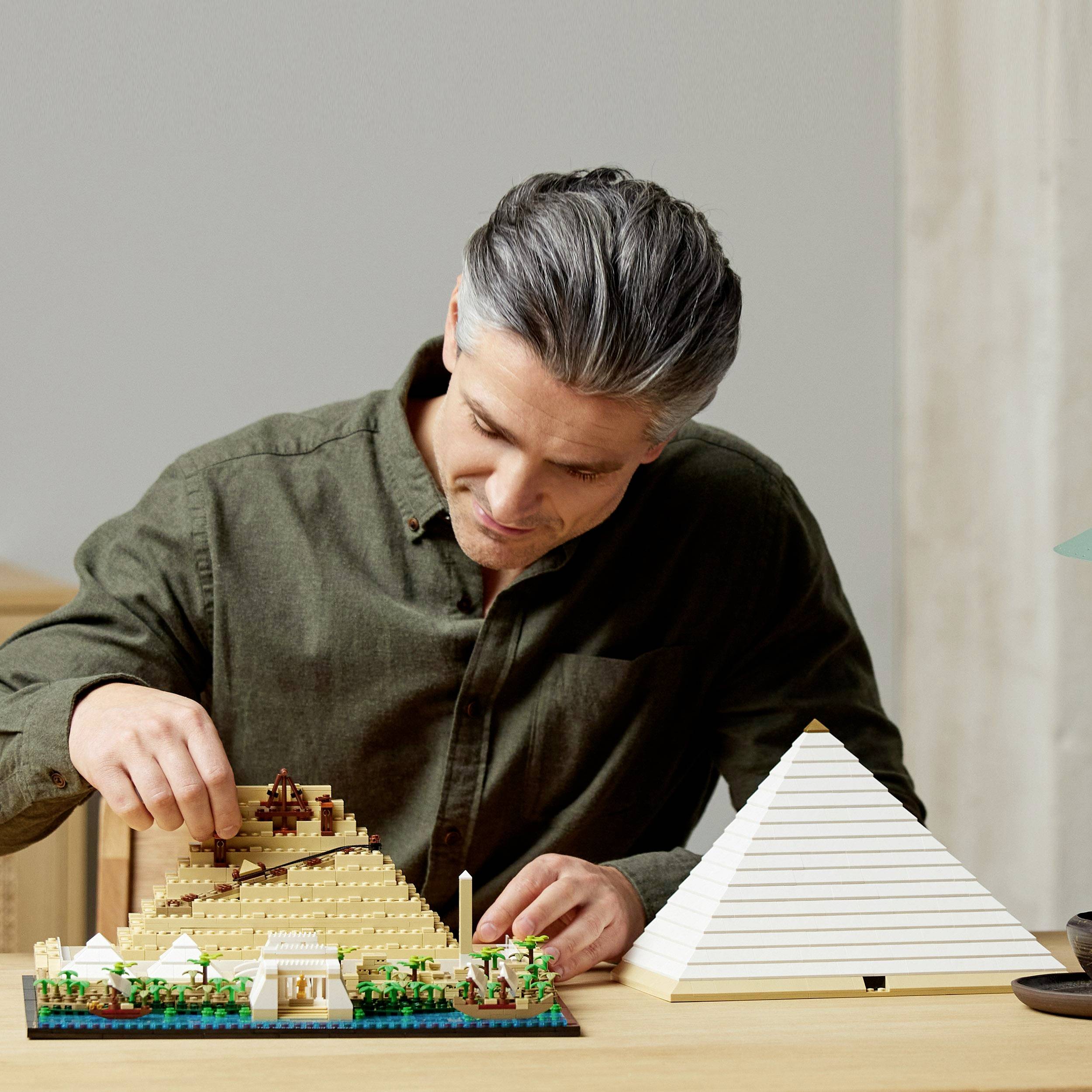 Een man bouwt een piramide van bouwstenen op een tafel, terwijl naast hem een voltooide witte modelpiramide staat.