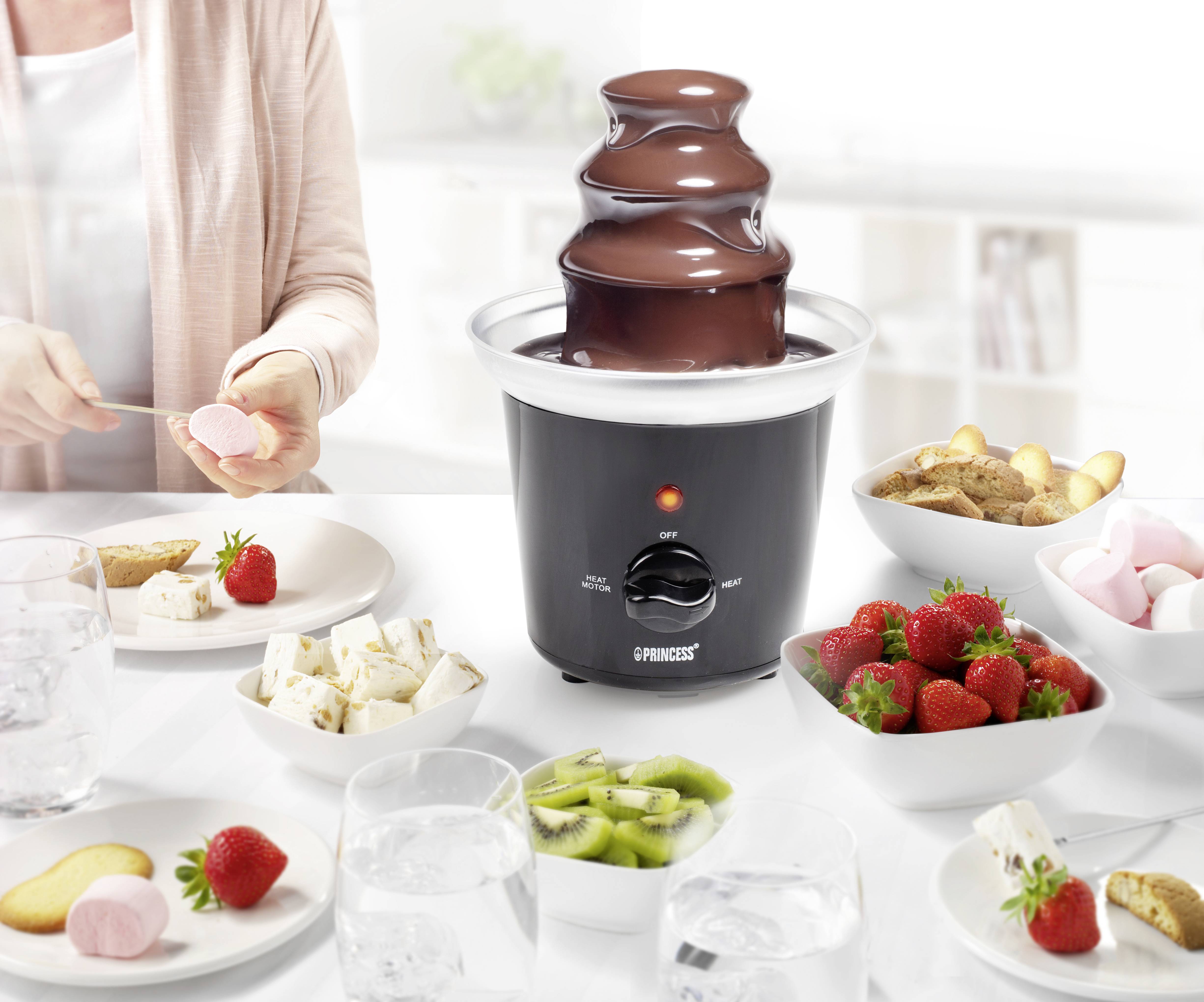 Chocoladefontein met stromende chocolade, omringd door aardbeien, kiwi, marshmallows en koekjes. Één persoon is bezig gerechten voor te bereiden.