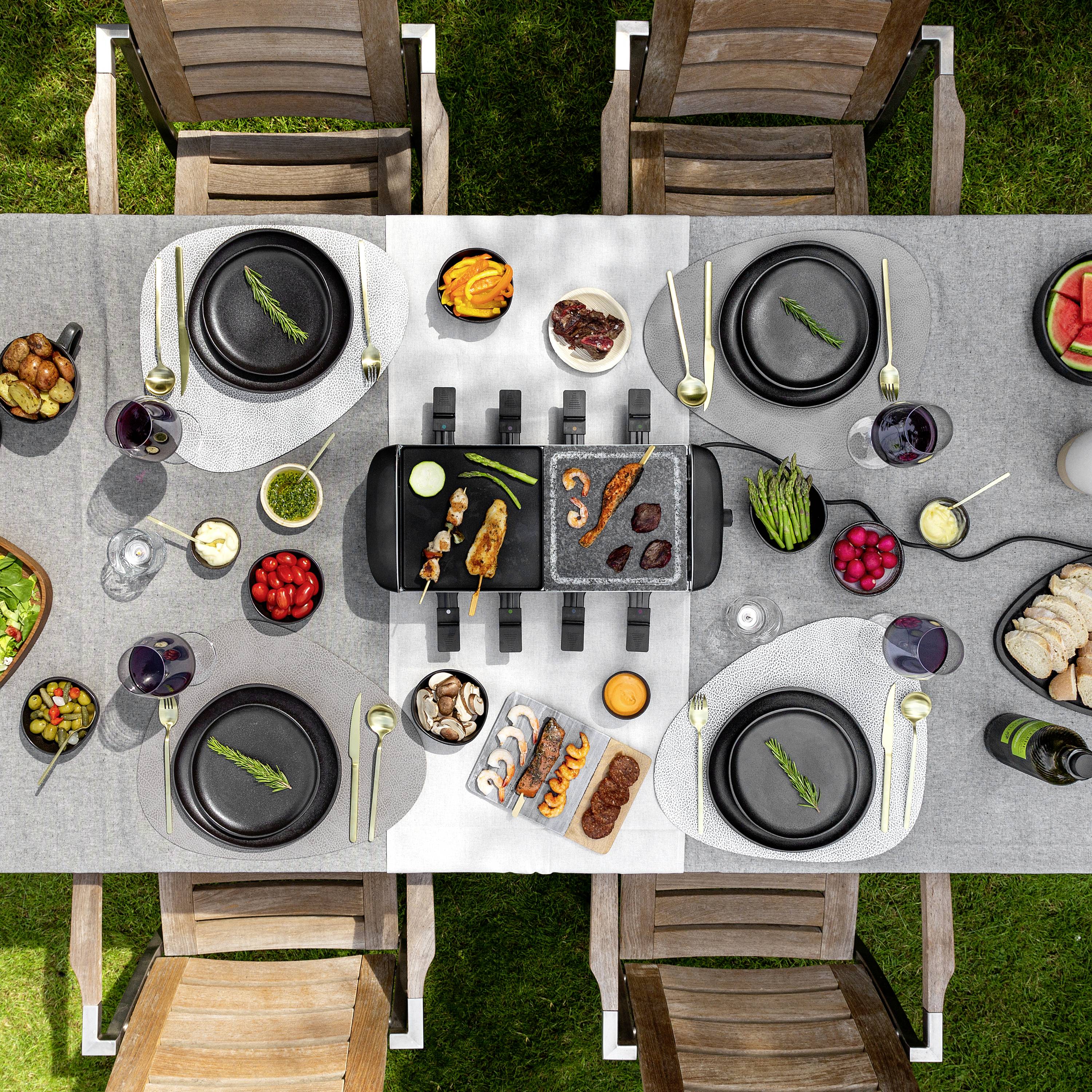 Een eettafel buiten is gedekt met een elektrische grill in het midden. Verschillende gerechten, salades, dranken en servies zijn te zien.