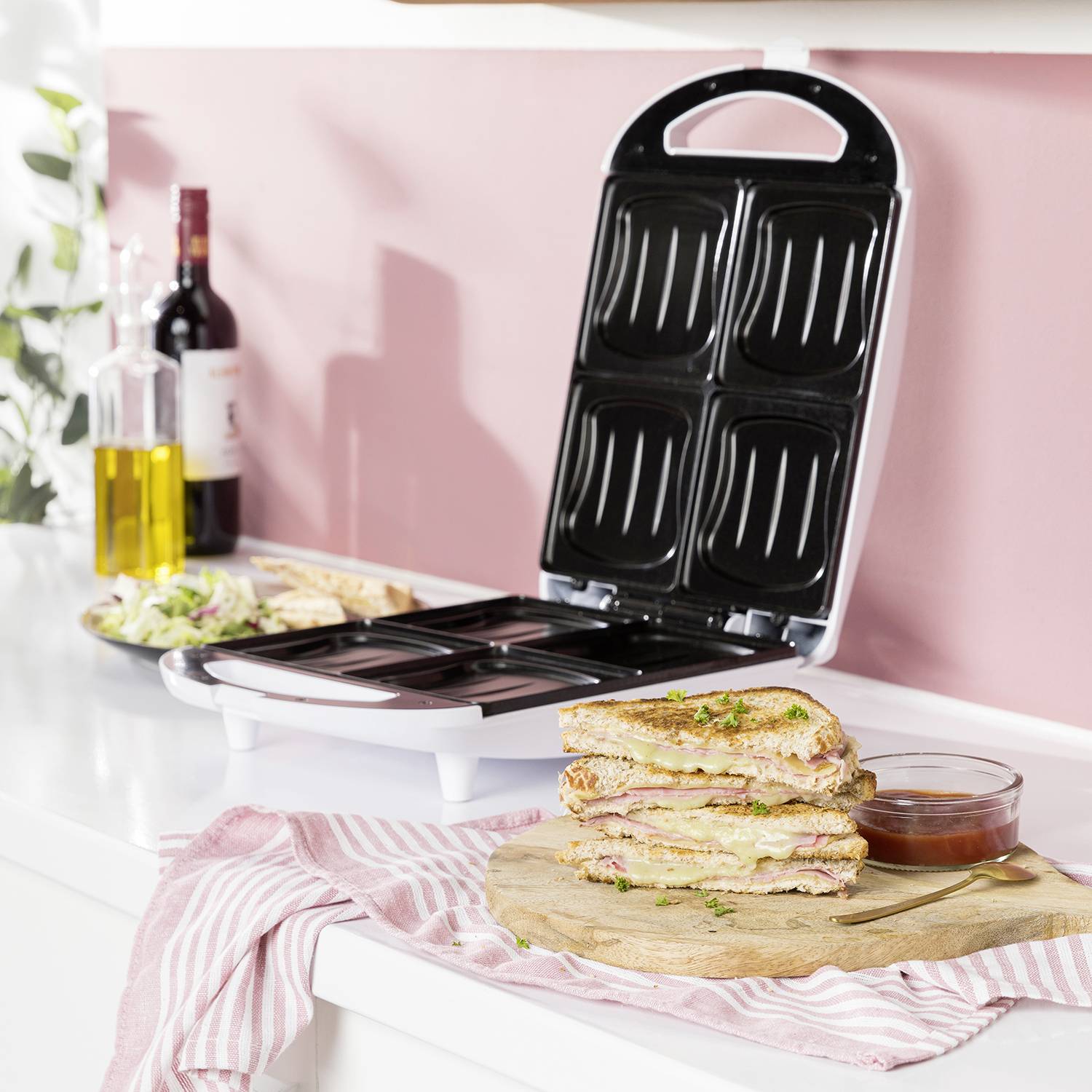 Een tosti-ijzer staat open op een aanrecht. Ernaast staat een bord met belegde broodjes en een schaaltje saus.