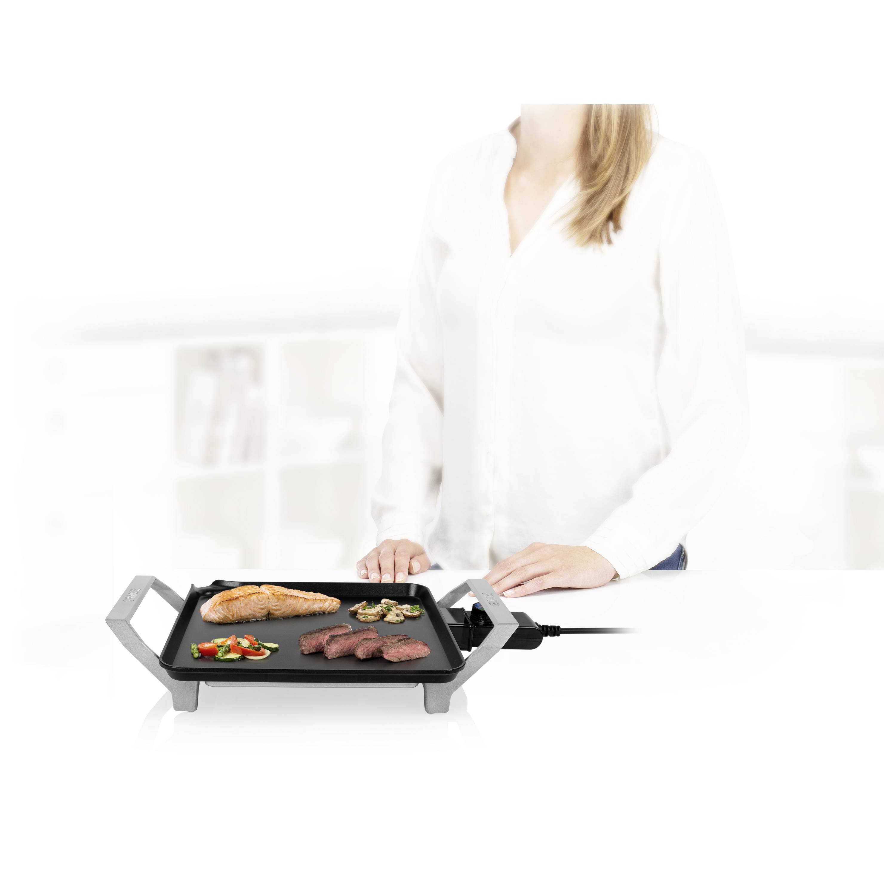 Een vrouw staat naast een tafelgrill met vlees, vis en groente erop, in een moderne keuken.