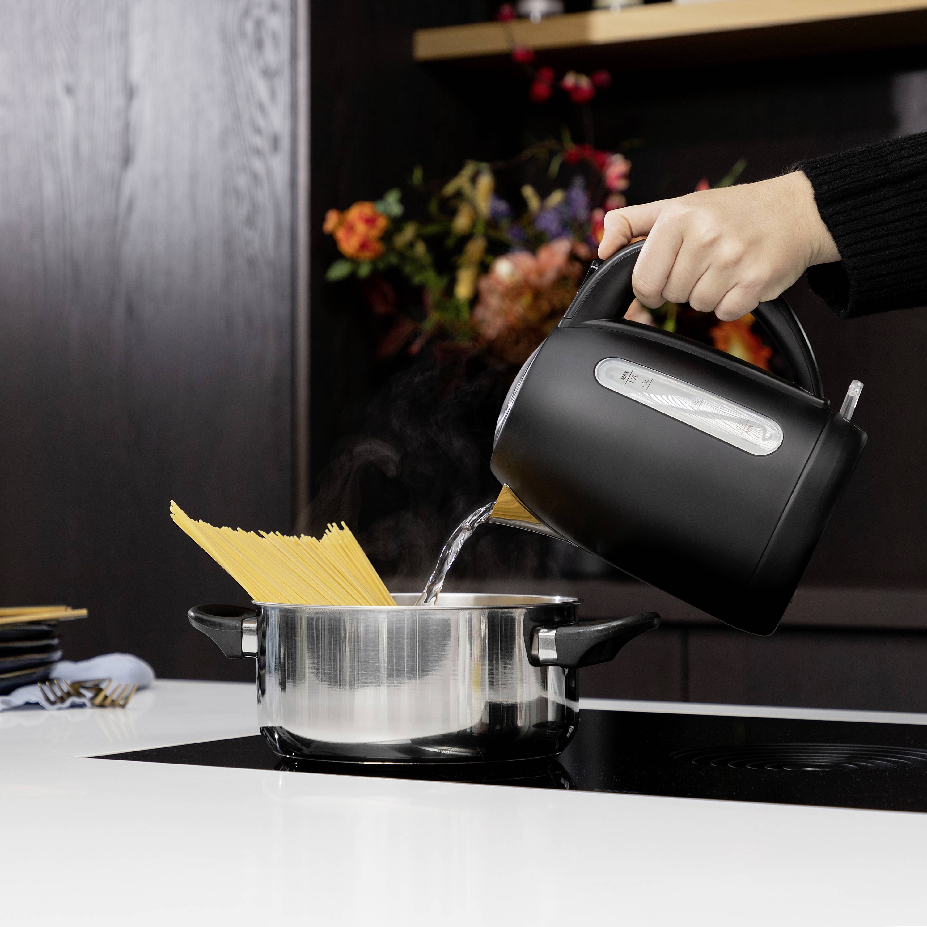 Een hand giet water uit een waterkoker in een pan met droge spaghetti om ze te koken. Op de achtergrond zijn bloemen te zien.