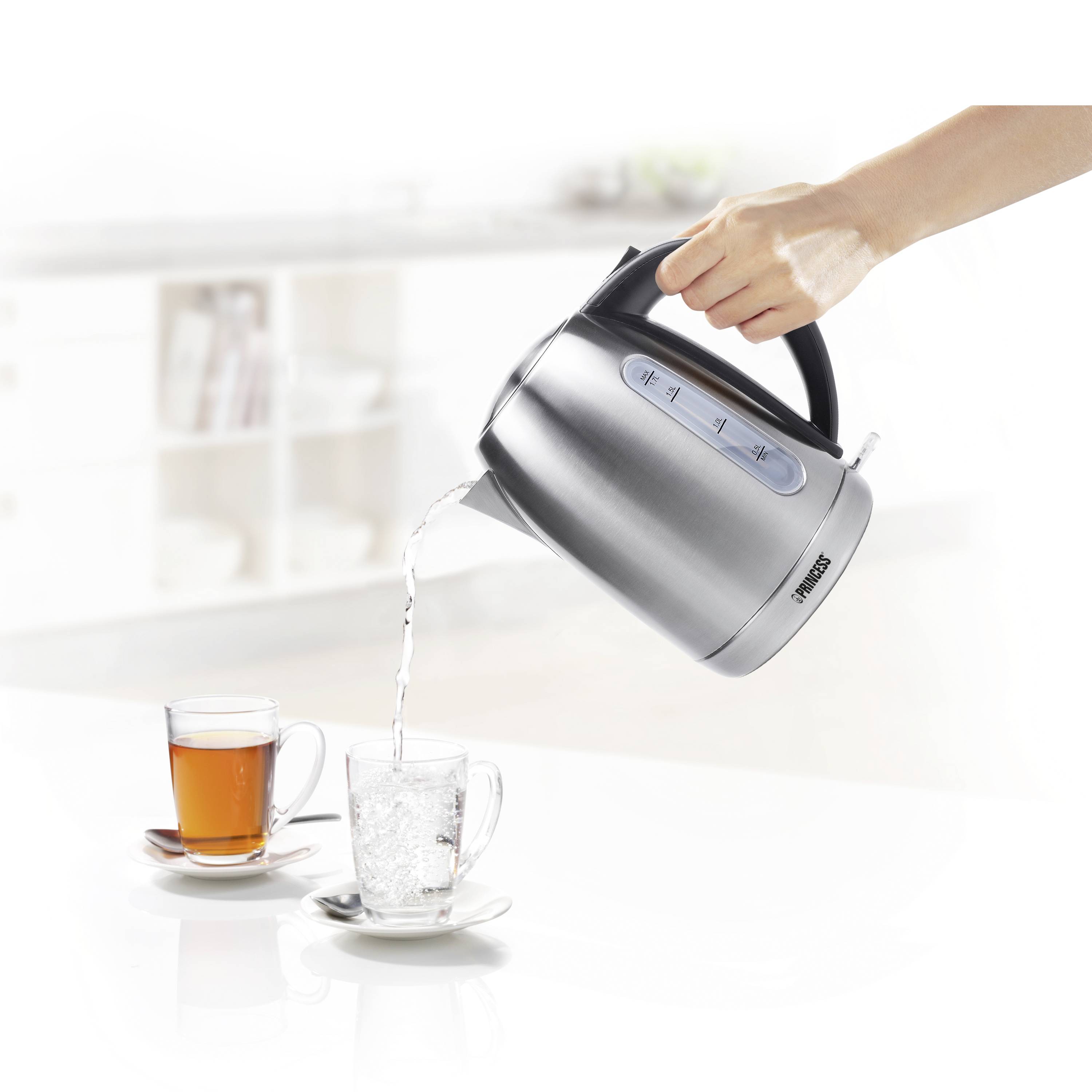 Een hand giet water uit een zilveren waterkoker in een kopje, naast een kopje thee op een witte tafel.