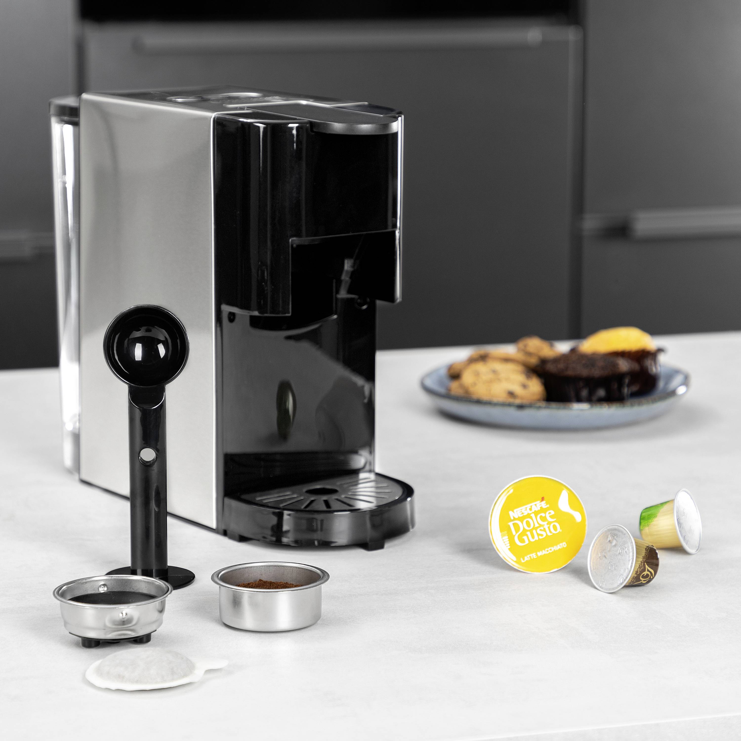 Koffiemachine op een wit oppervlak, omringd door koffiecapsules en een koffielepel. Erachter staat een bord met koekjes en muffins.