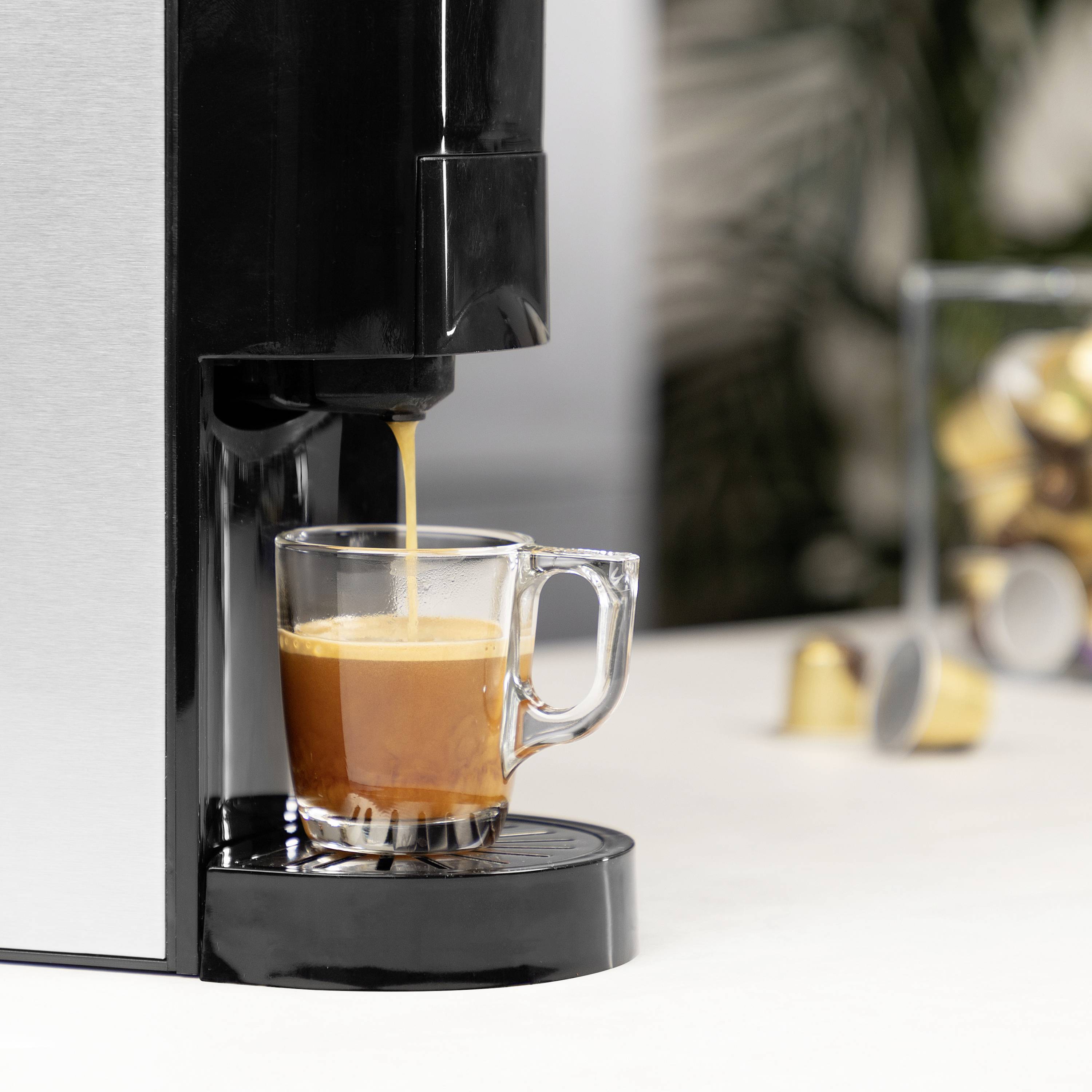 Een koffiemachine zet verse espresso in een glazen kopje. Op de achtergrond zijn onscherpe koffiecapsules te zien.