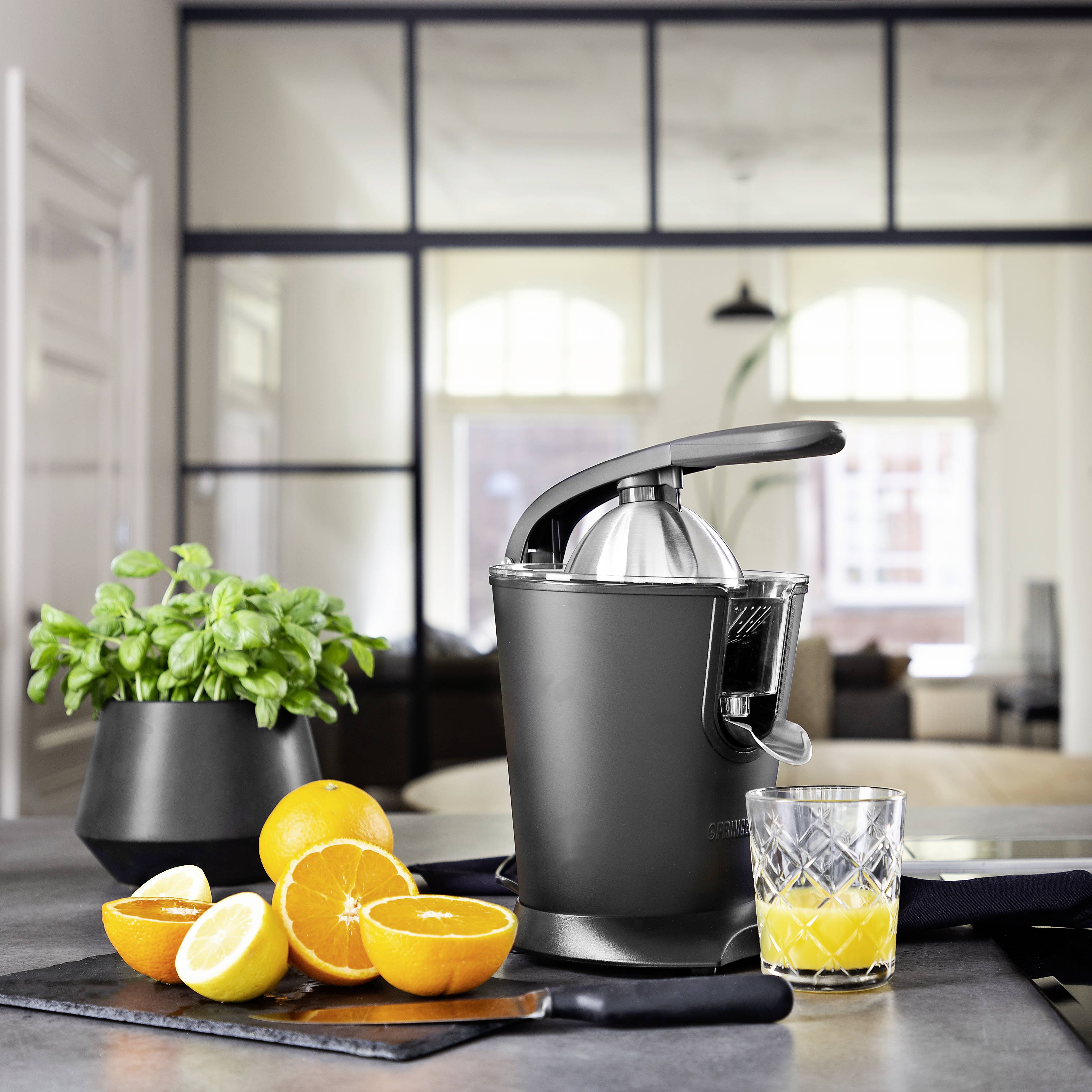 Elektrische citruspers op keukentafel naast een basilicumplant en gesneden sinaasappels. Glas vol sap op de voorgrond. Lichte, moderne keuken.