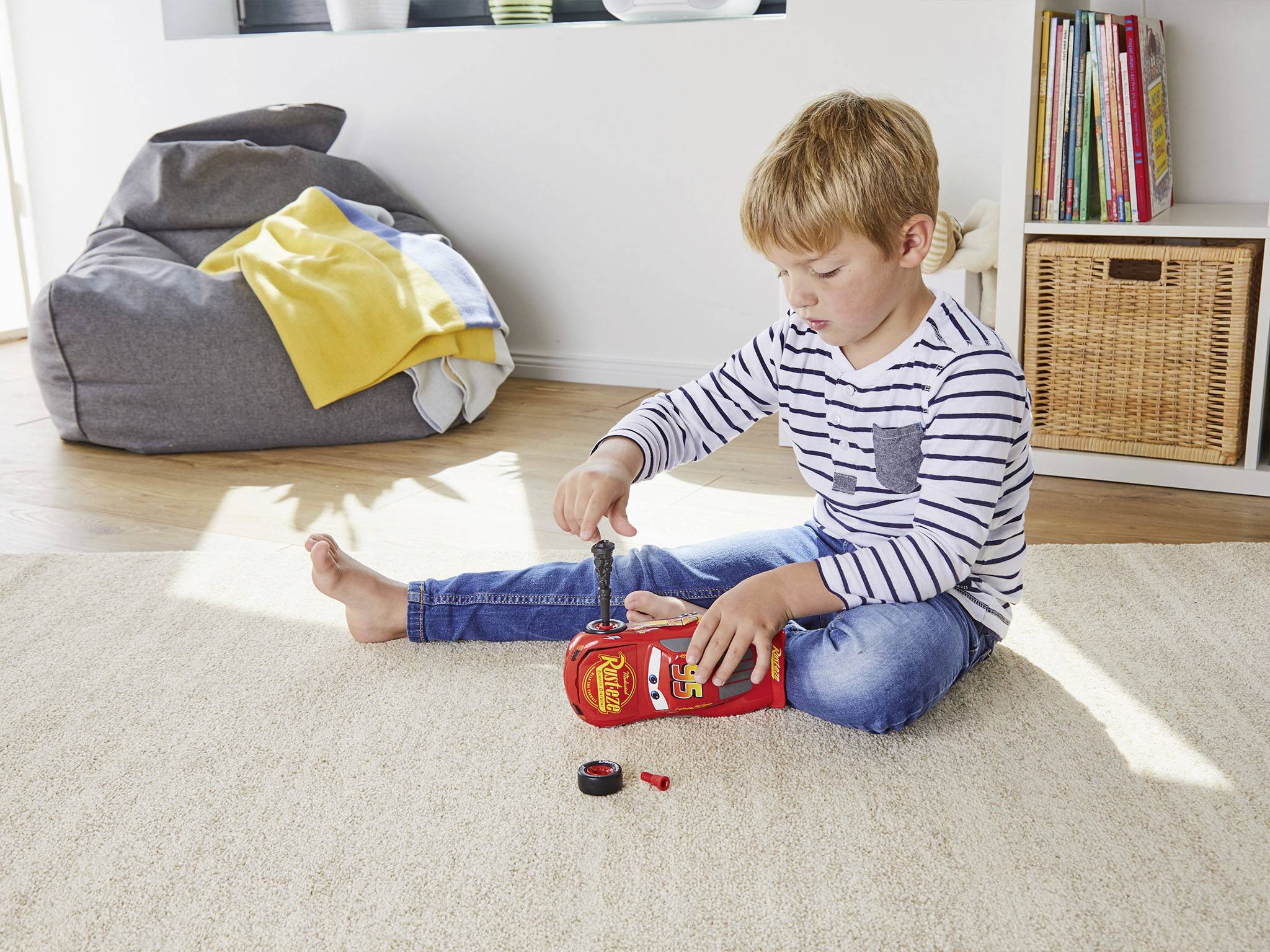 Een jongen zit op een vloerkleed en speelt met een speelgoedauto. Op de achtergrond zijn een kussen en een boekenkast te zien.