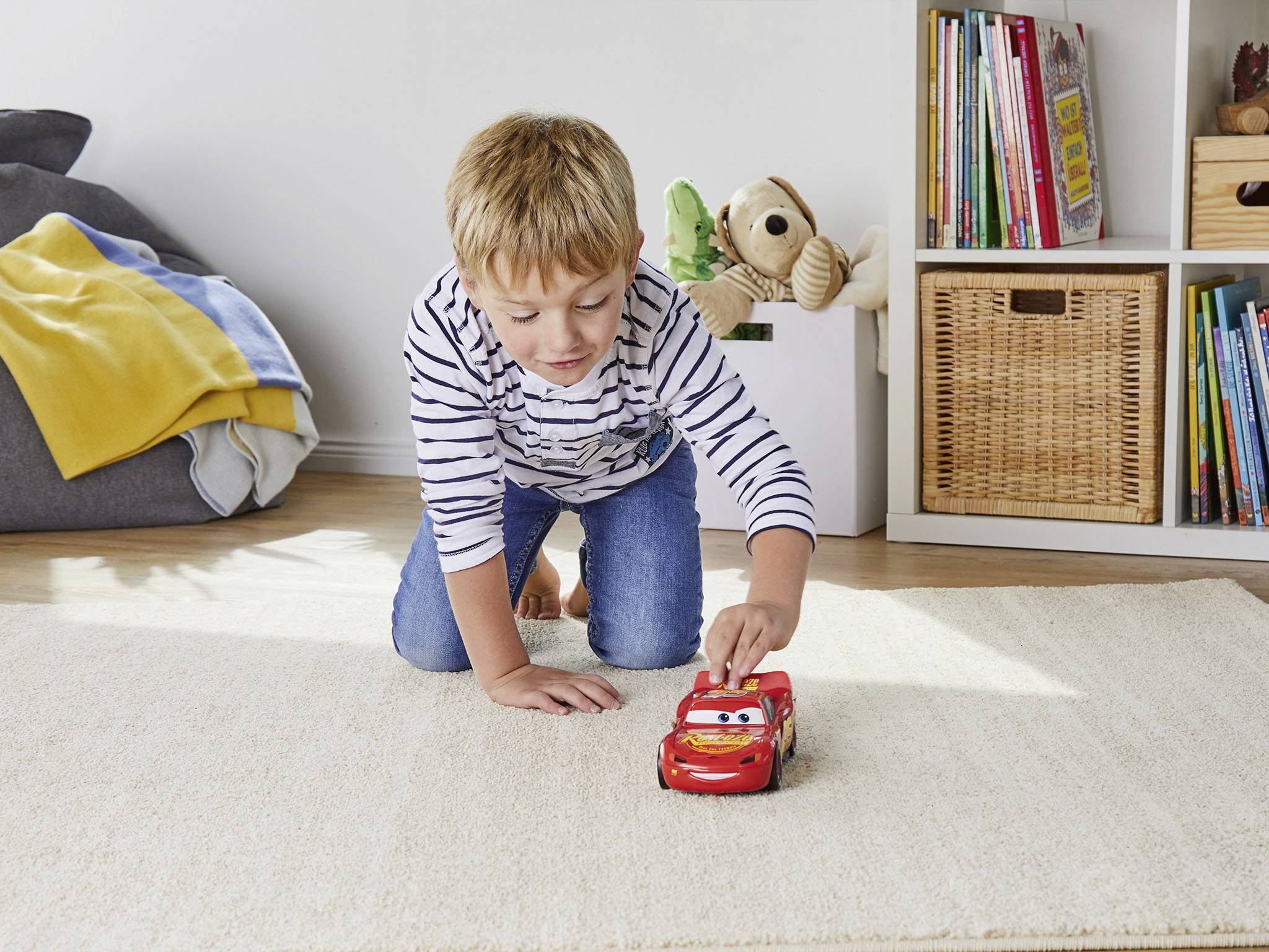 Een jongen speelt op een vloerkleed met een rode speelgoedauto in een kinderkamer. Op de achtergrond boeken en een knuffeldier.
