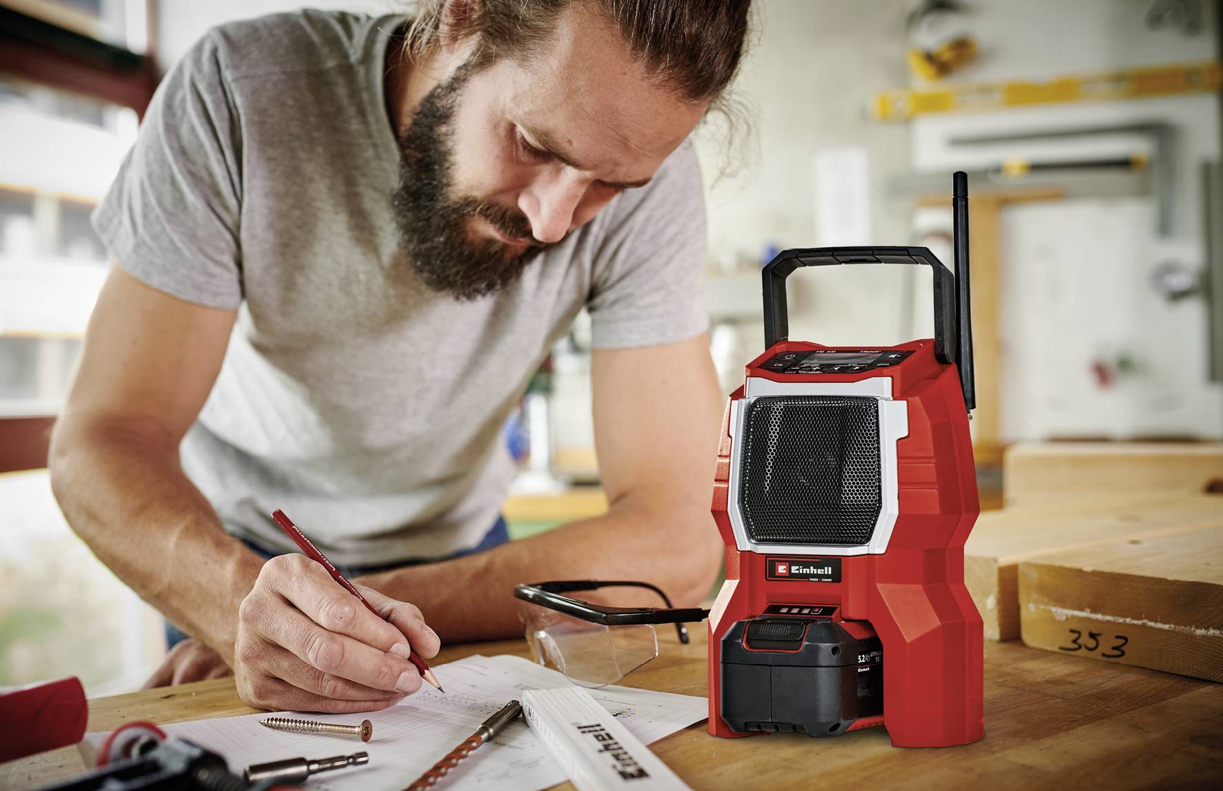 Een man met een baard maakt aantekeningen op papier in een werkplaats. Op tafel staat een rood bouwradio.