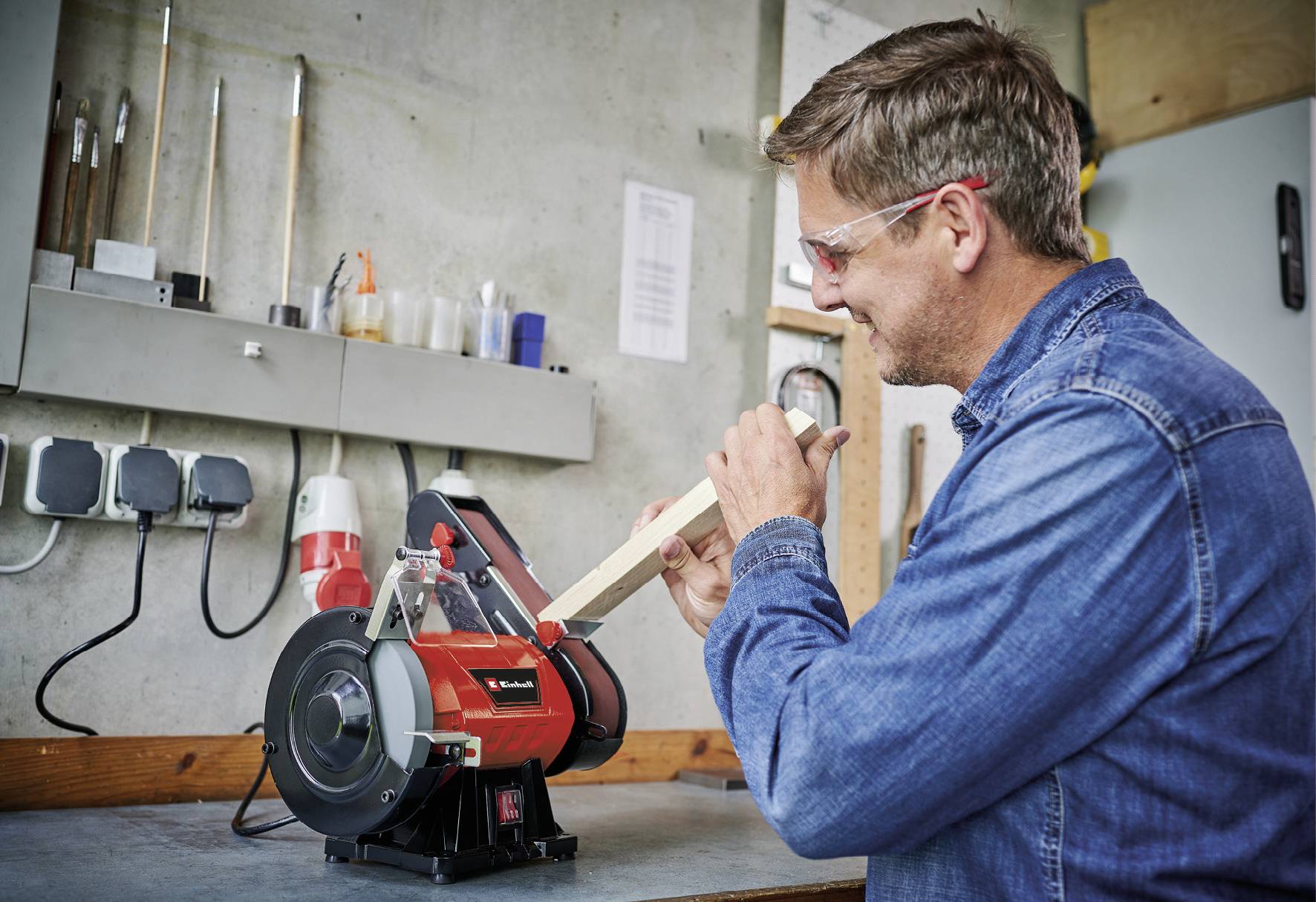 Een man slijpt een stuk hout met een bandschuurmachine in een werkplaats. Hij draagt een veiligheidsbril en een spijkerhemd.