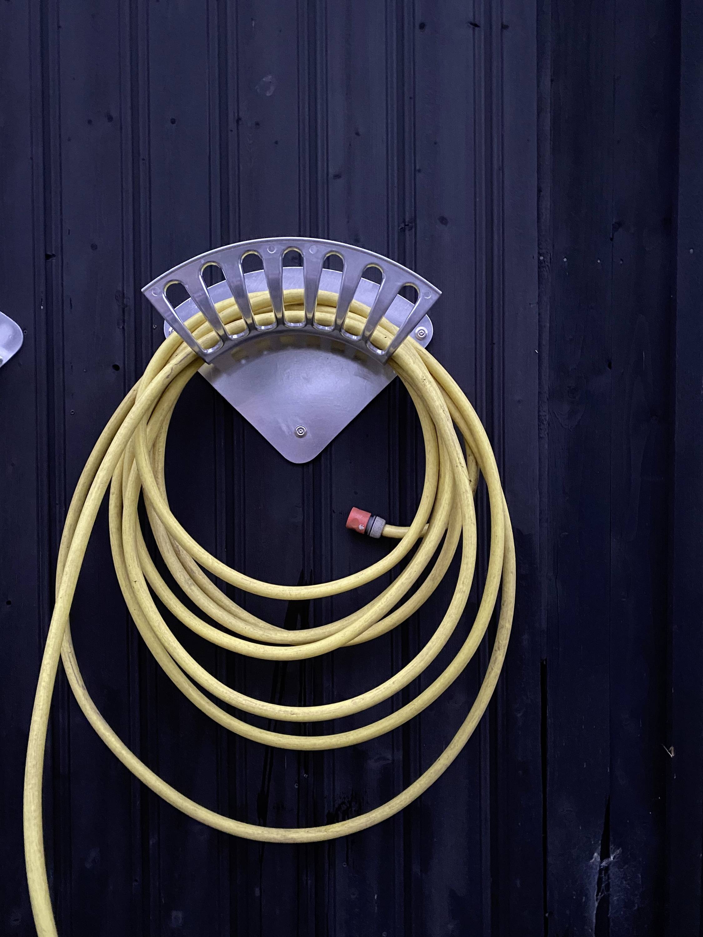 Een gele tuinslang is netjes opgerold aan een metalen wandhouder aan een zwarte muur.