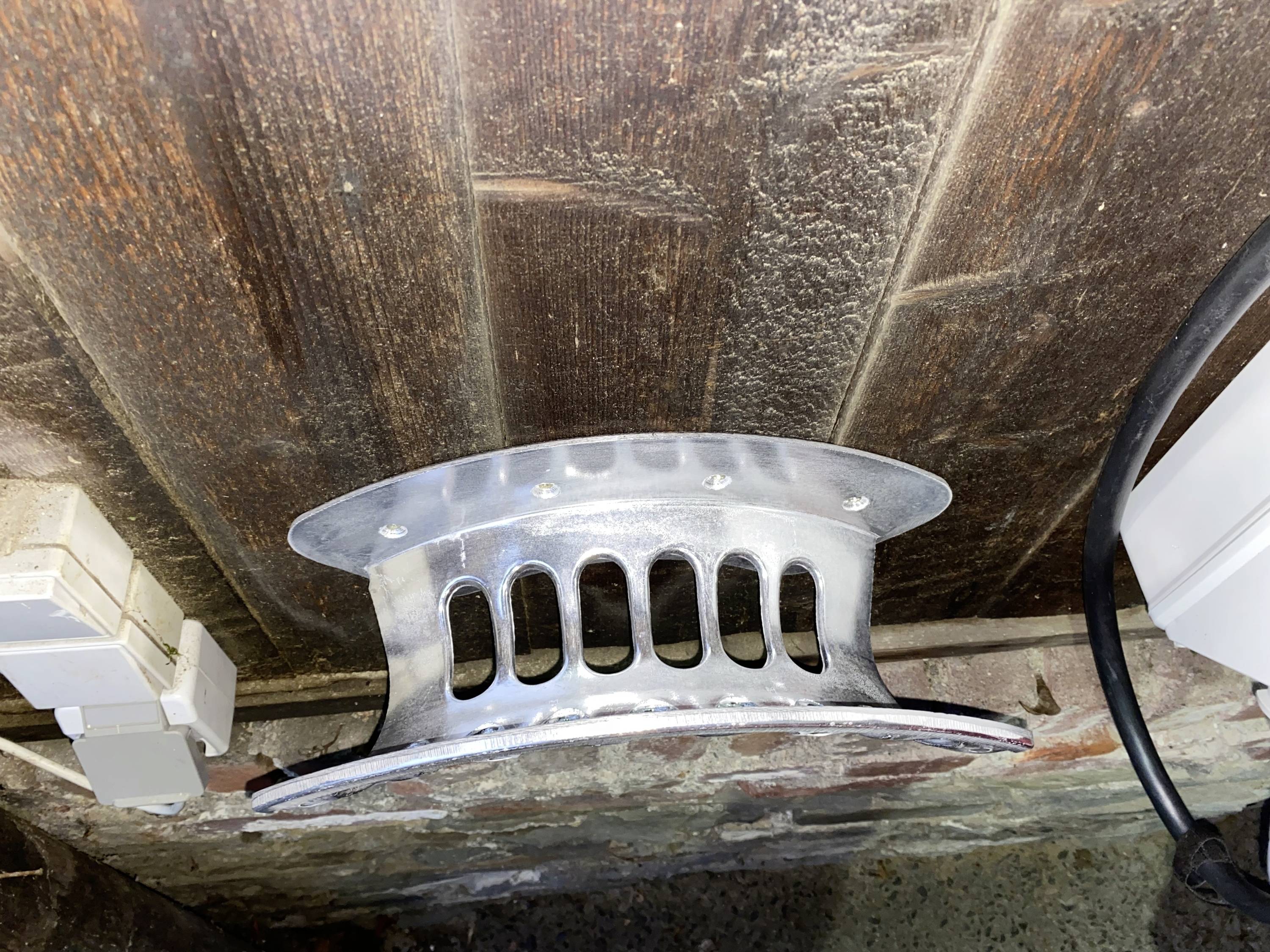 Metalen stuk gemonteerd aan een houten muur, boven elektrische aansluitingen. Structuren achter en rechts zichtbaar.