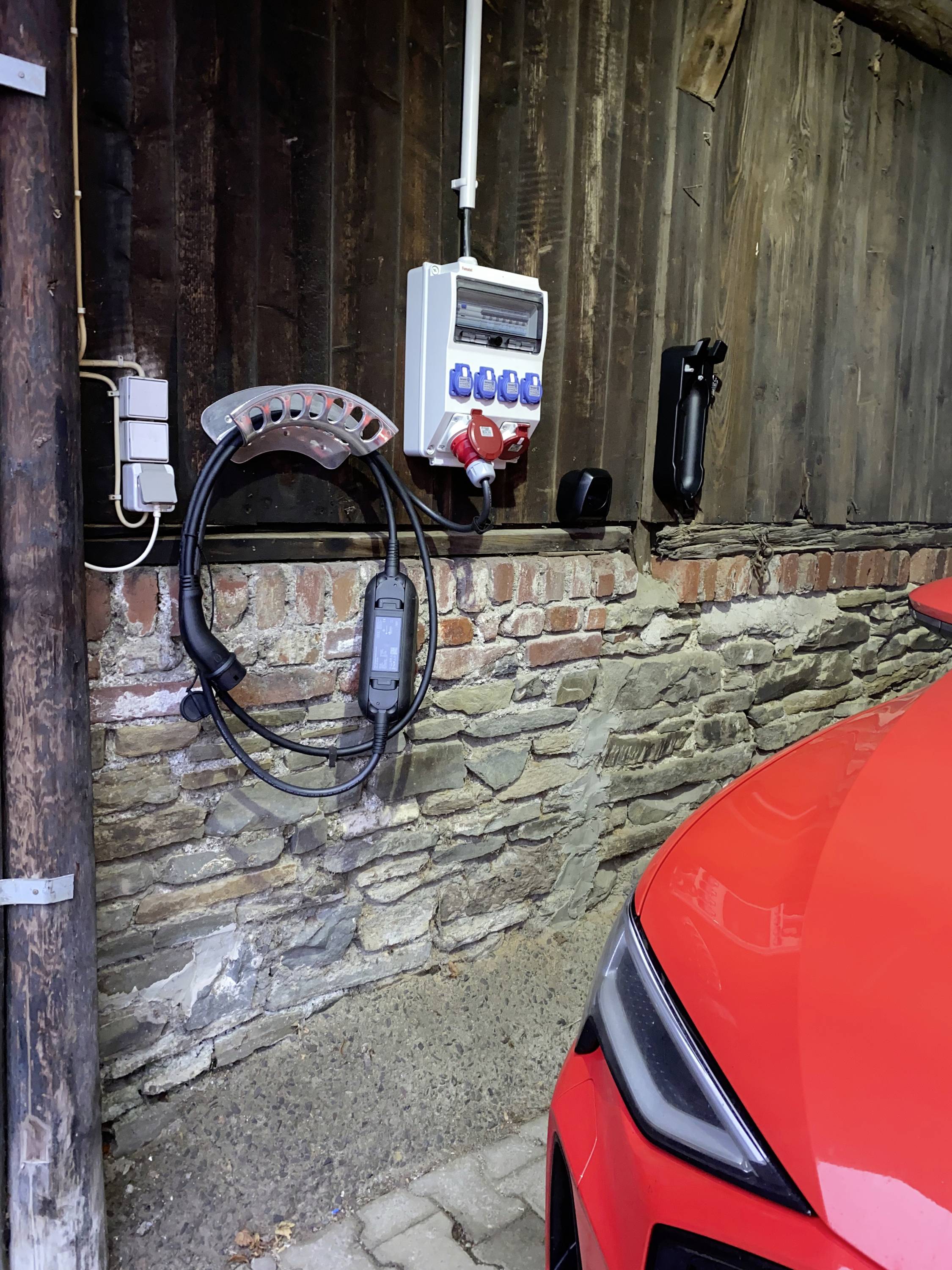 De voorkant van een rode auto naast een muur met een laadvoorziening. Aan de muur zijn een stopcontact en een laadkabel bevestigd.